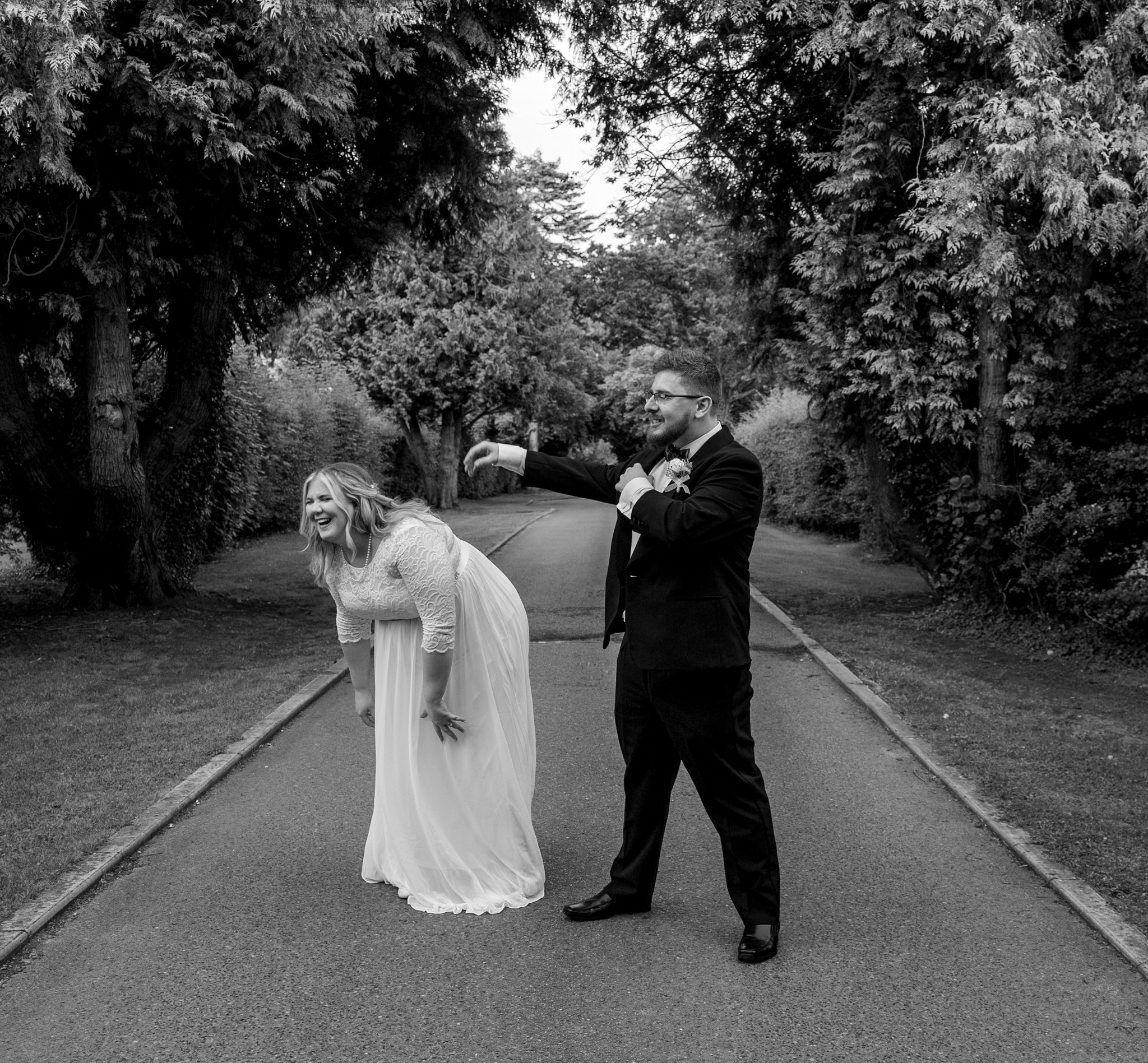 A bride and groom in a playful pose on a tree-lined path.