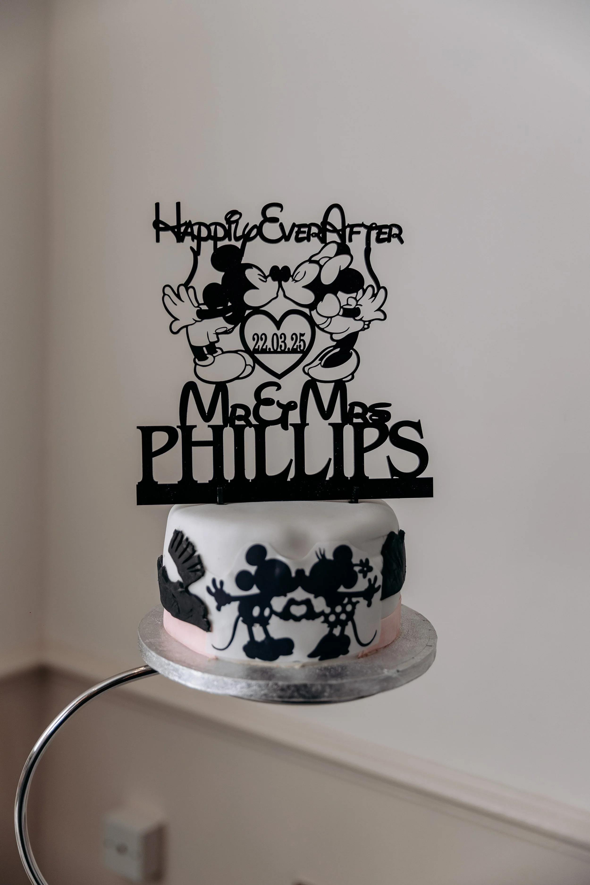 A wedding cake decorated with Mickey and Minnie Mouse themed cake toppers and cutouts, including a top topper with the text 'Happy Ever After,' a date, and the names Mr. & Mrs. Phillips.