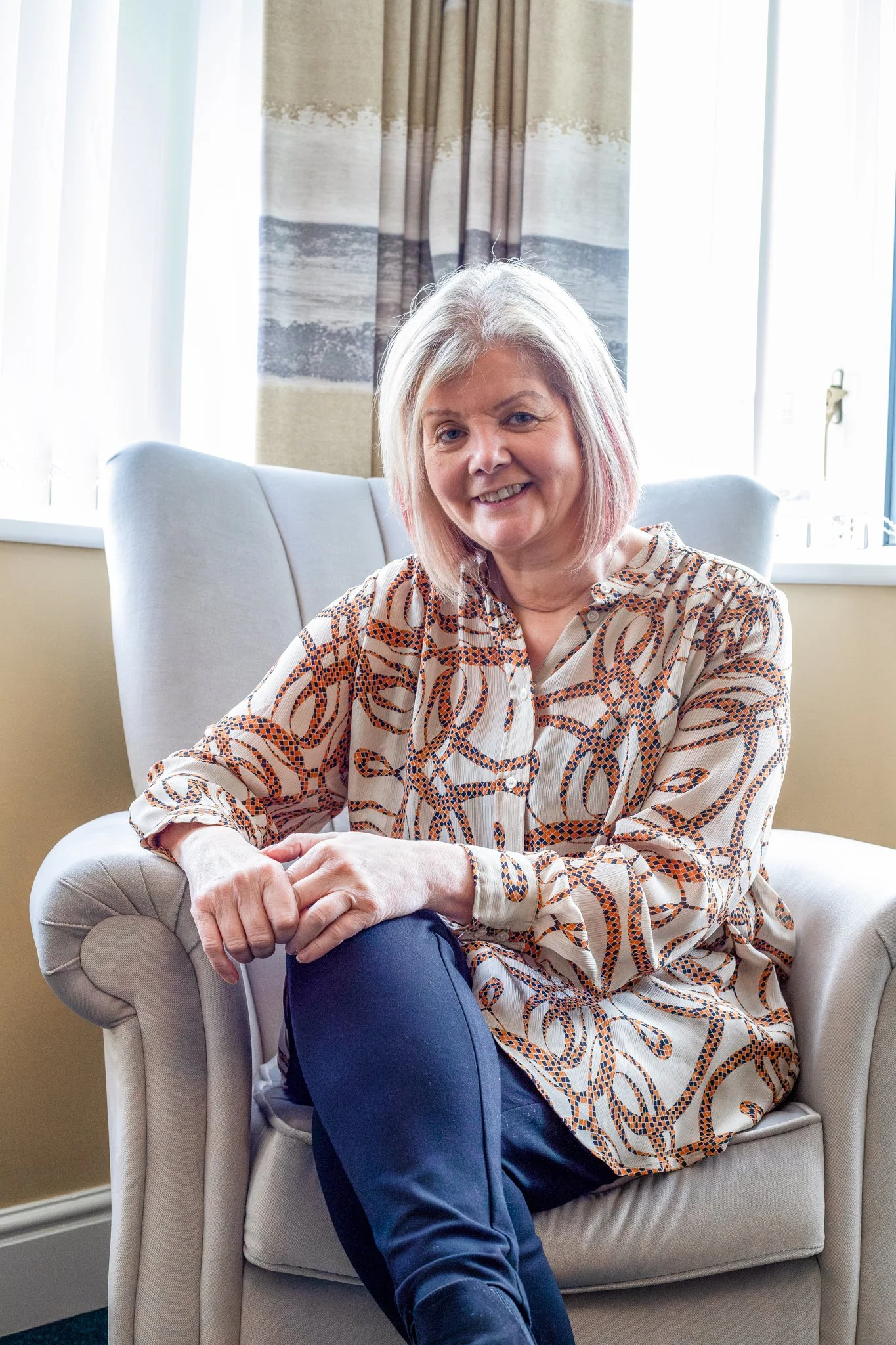 A middle-aged woman with blonde hair and a patterned shirt, sitting on a beige armchair in front of a window with curtains, smiling at the camera.