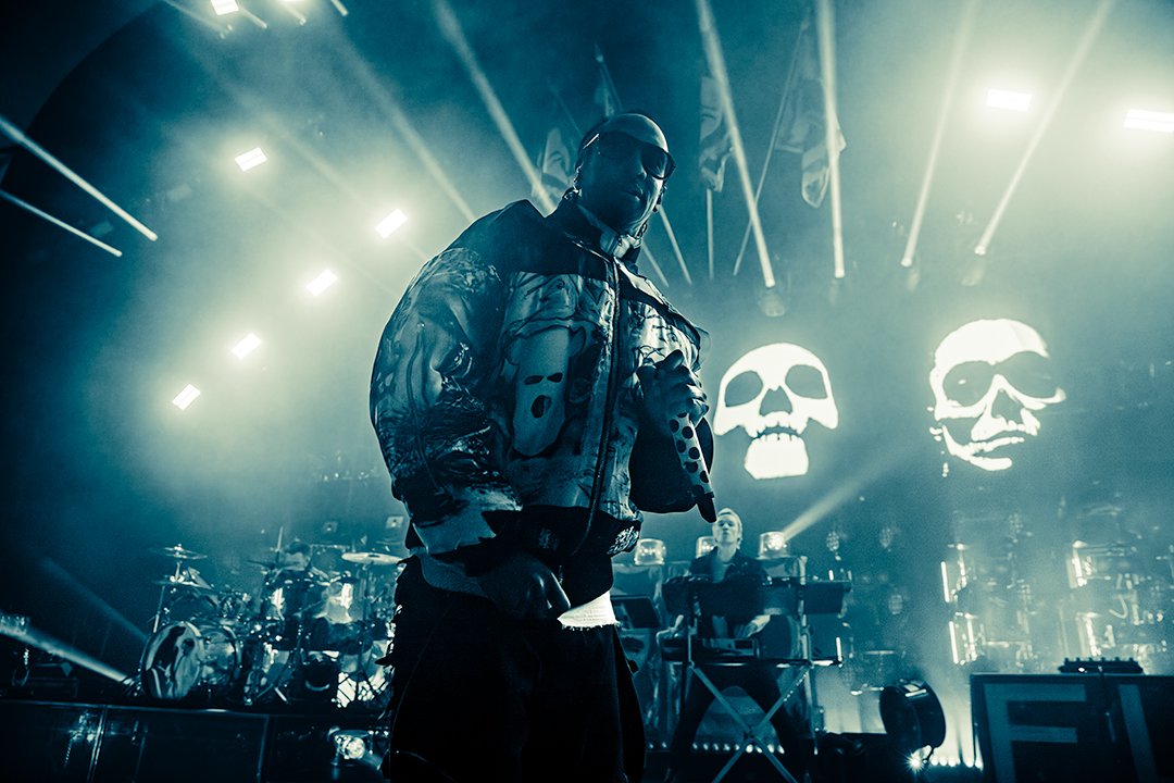 Musical performer on stage holding microphone during concert with LED skull visuals in background and stage lights.