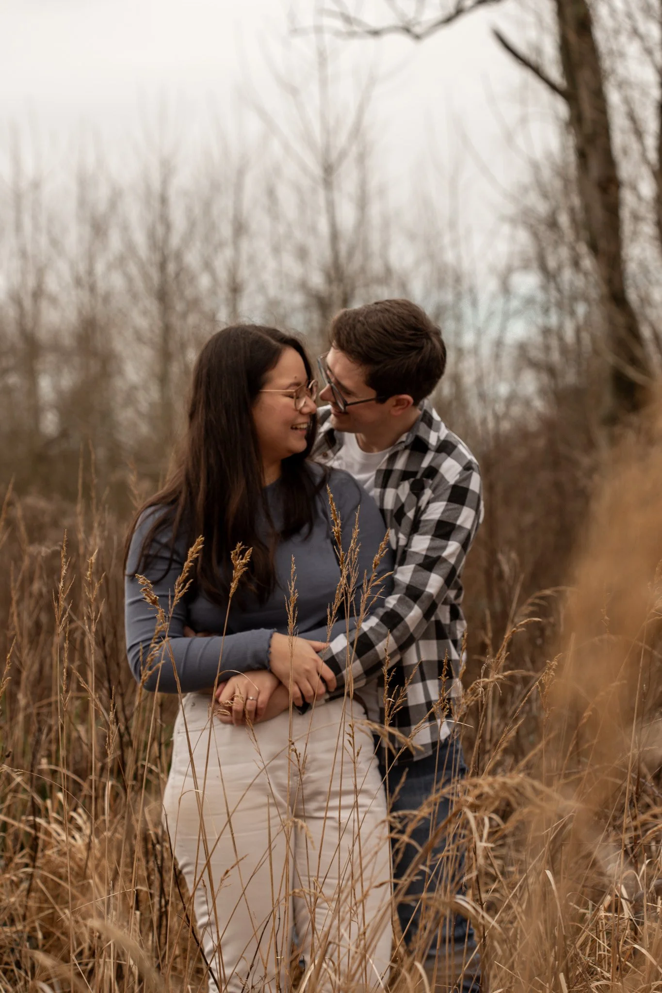A couple embracing and smiling in a field of tall, dry grass with leafless trees in the background on a cloudy day.