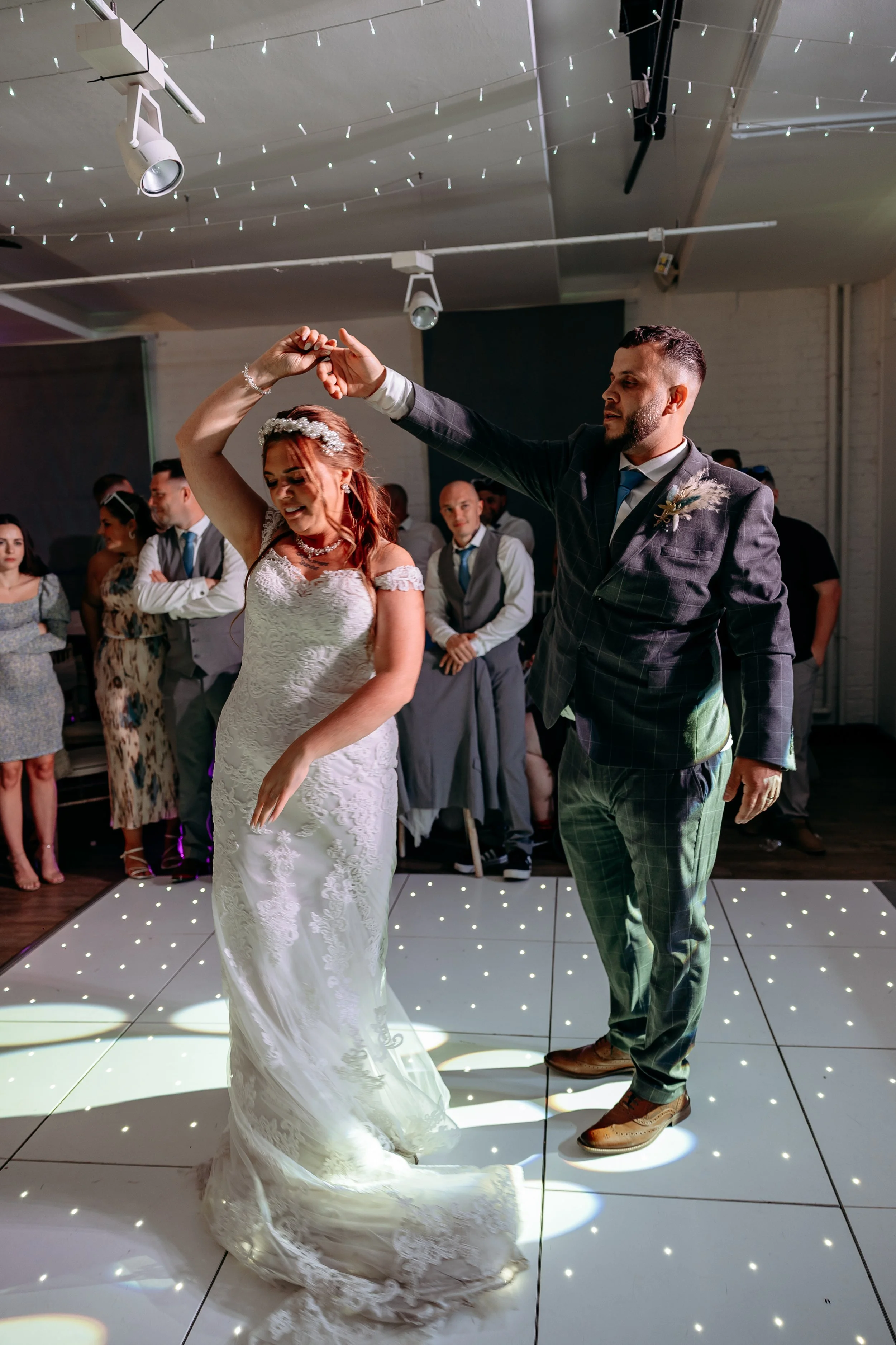 Bride and groom dancing on a wedding reception dance floor, surrounded by guests, with decorative lights overhead.