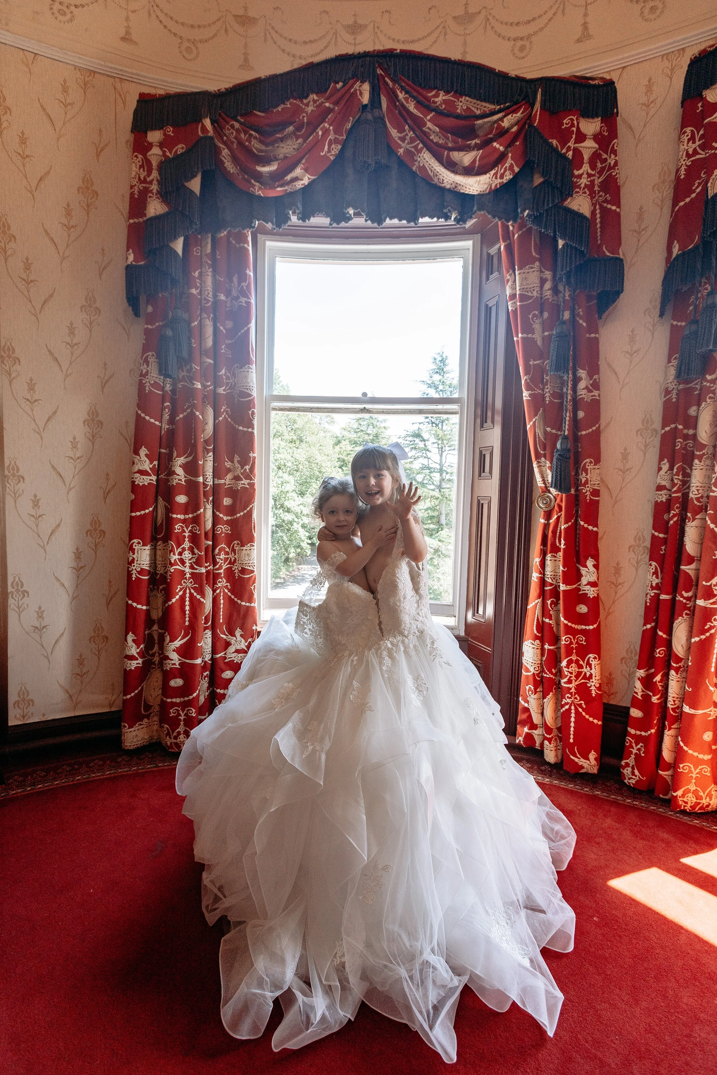 Two young girls in wedding dresses sitting on a window ledge, smiling and waving, inside a room with red and gold patterned curtains and wallpaper.