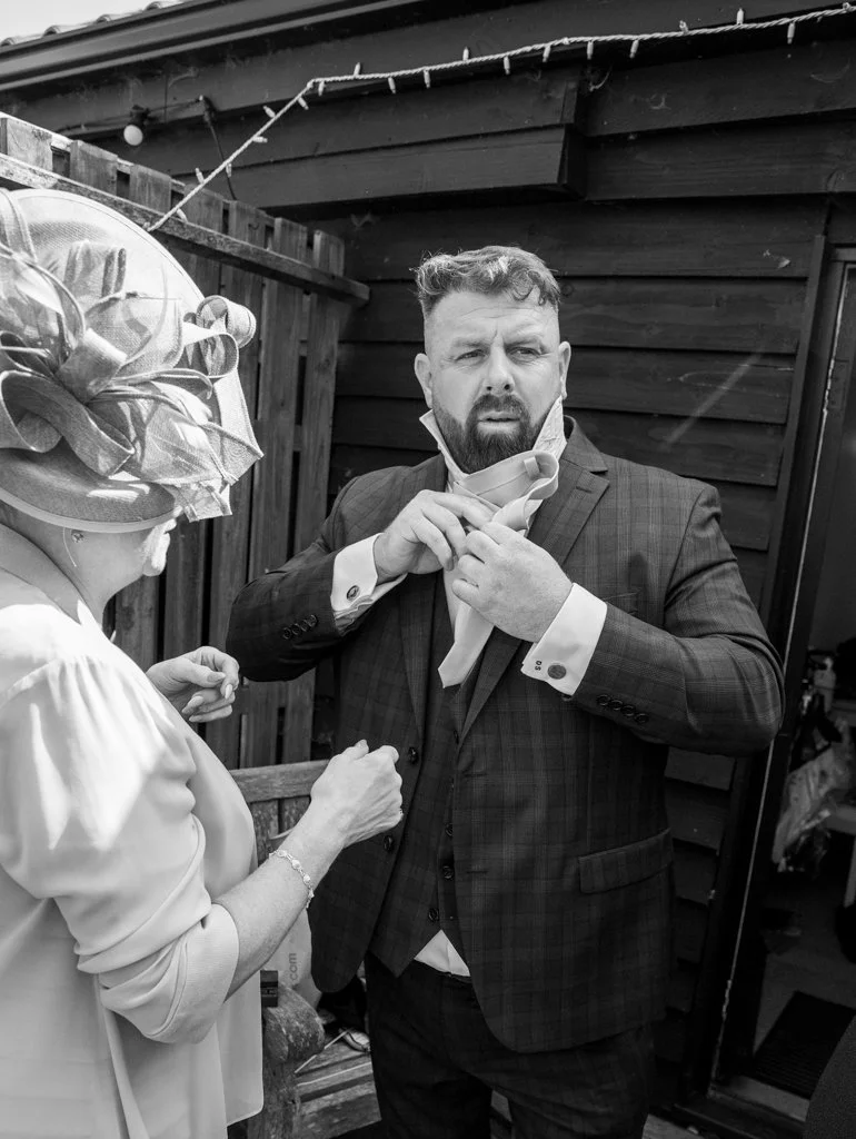 A man in a suit adjusts his necktie while standing outside next to an older woman wearing a large fascinator hat.