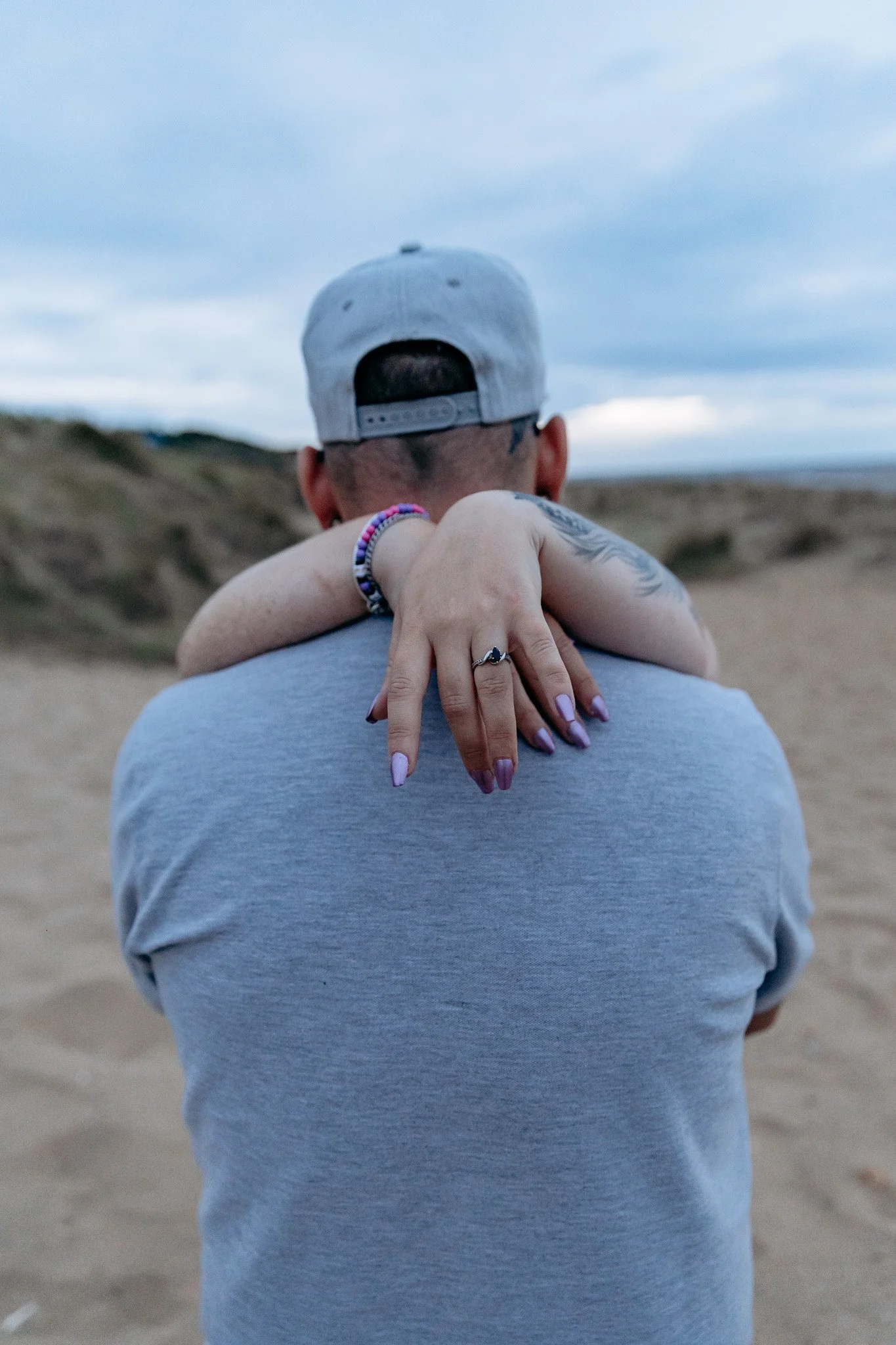 A person wearing a gray baseball cap and a gray T-shirt stands on a sandy beach with hills in the background. They are holding a woman in a hug, whose arms and hand with a ring are visible around their neck.