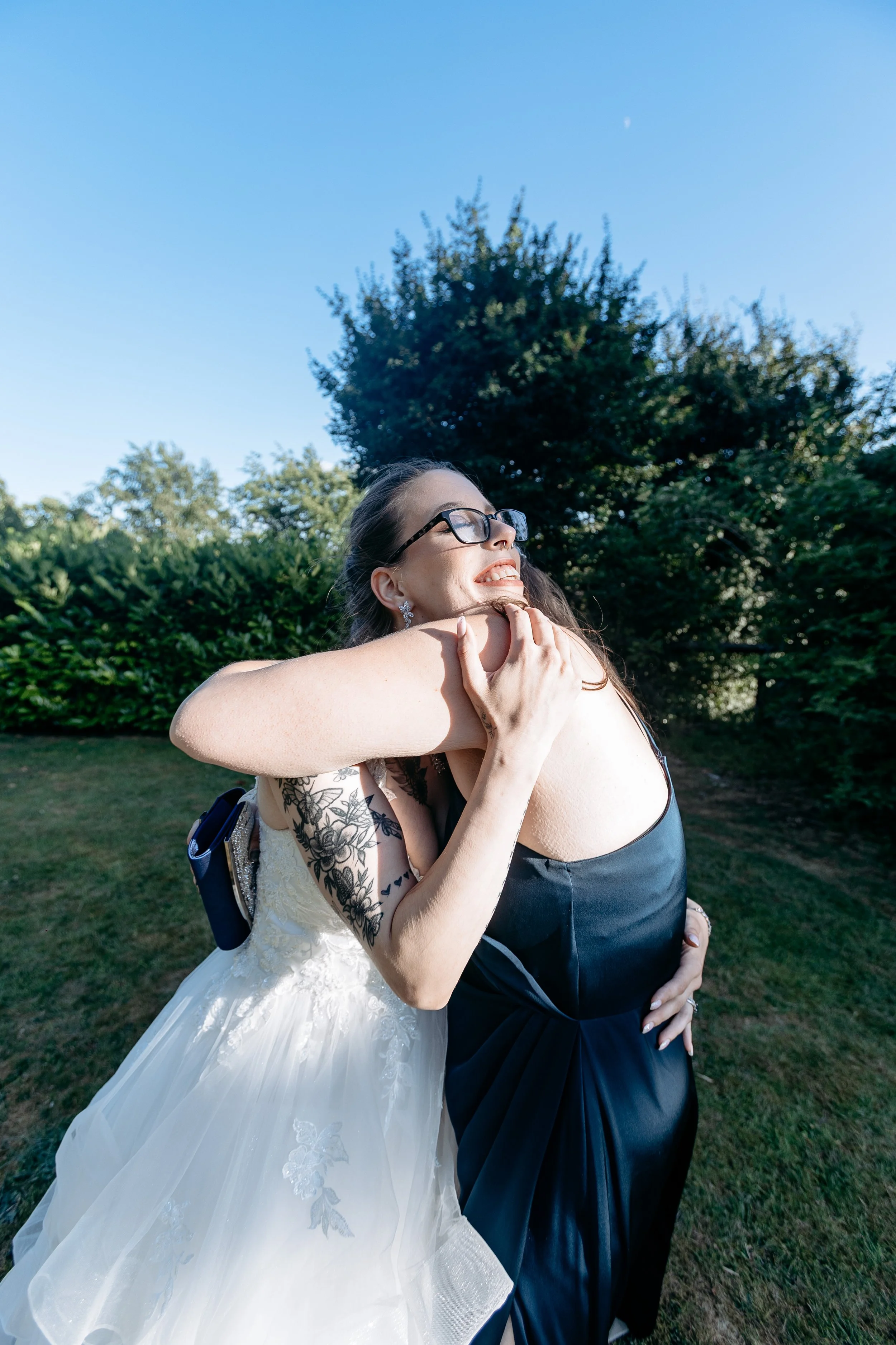 A woman in a wedding dress hugging another woman in a black dress outdoors on a sunny day with blue sky and trees in the background.