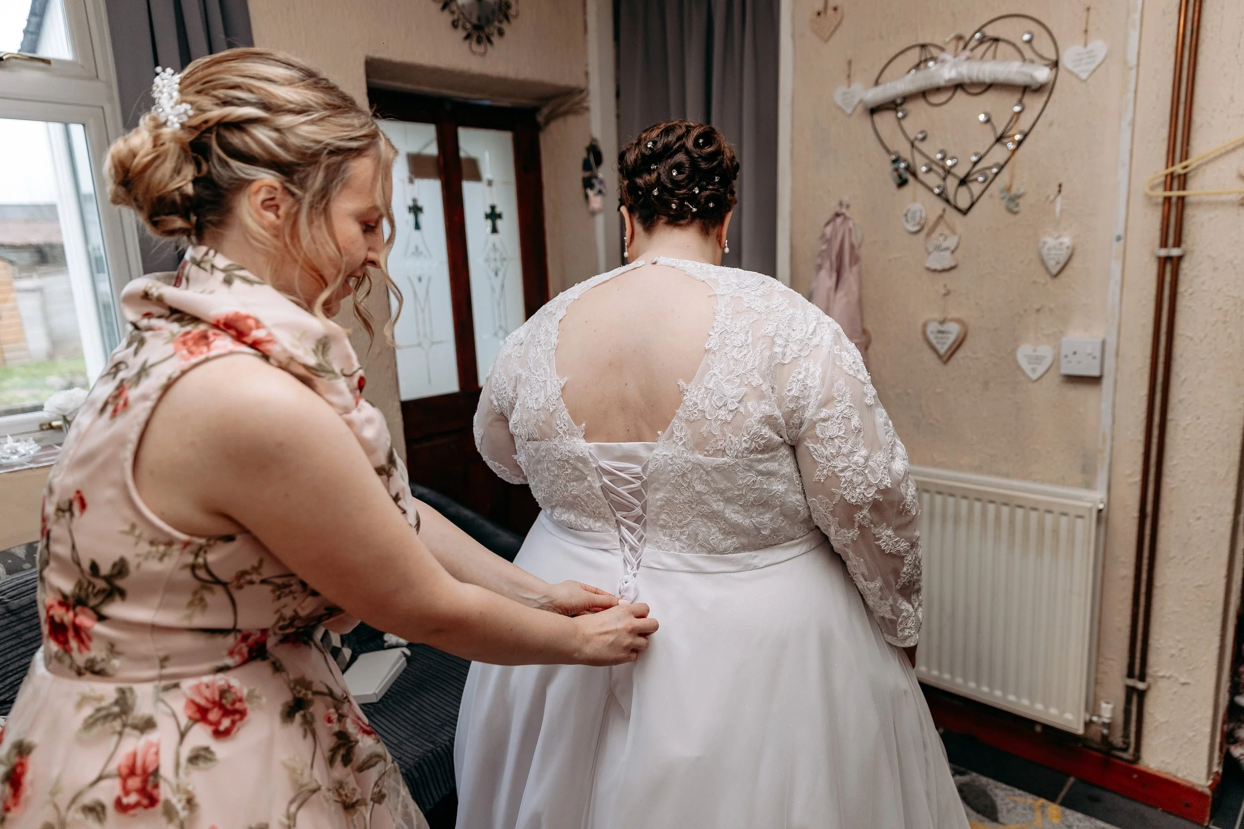 A woman in a floral dress helps another woman in a white bridal gown lace up the back of her dress in a decorated room.