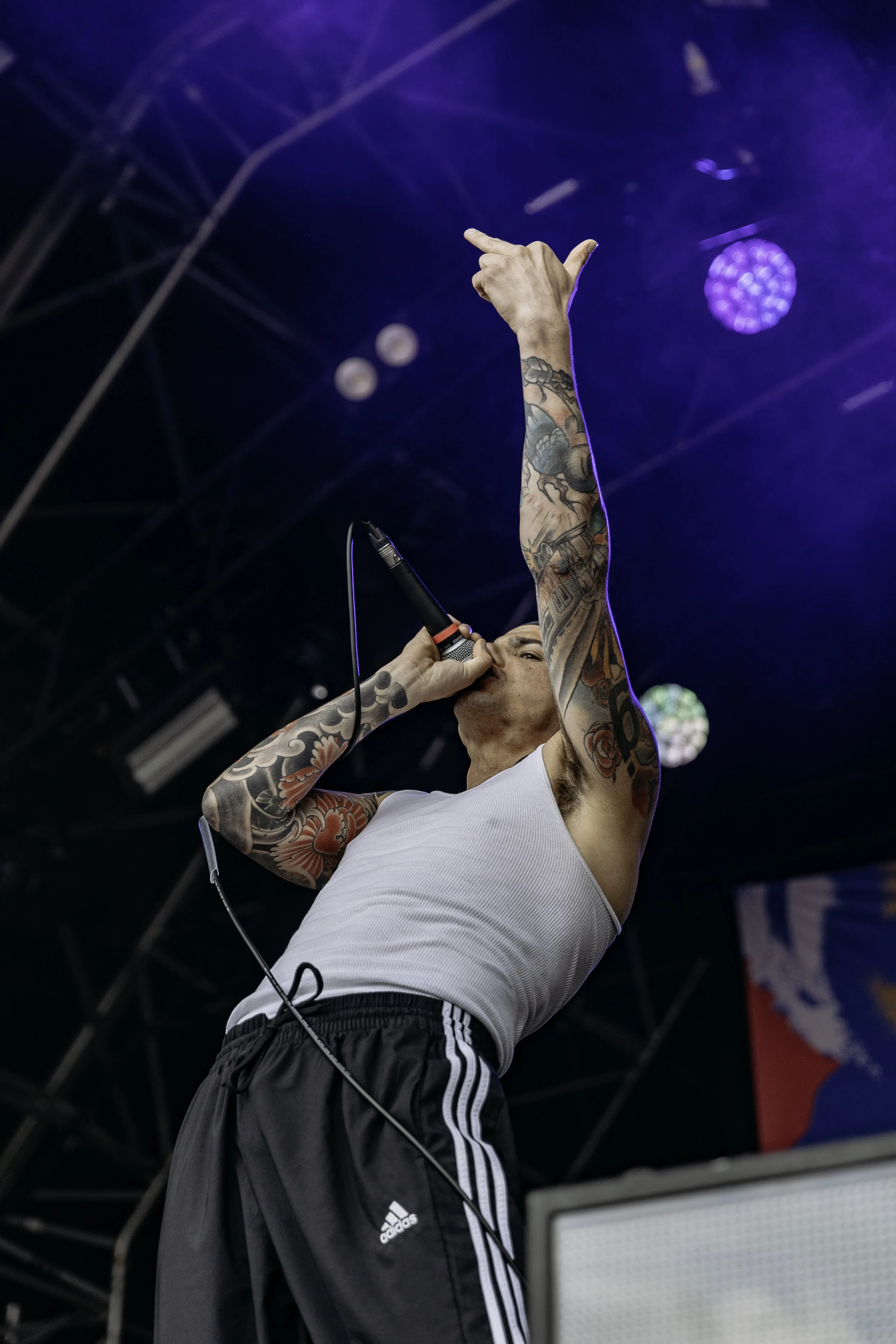 A tattooed man singing into a microphone on stage, wearing a white tank top and black Adidas sweatpants.