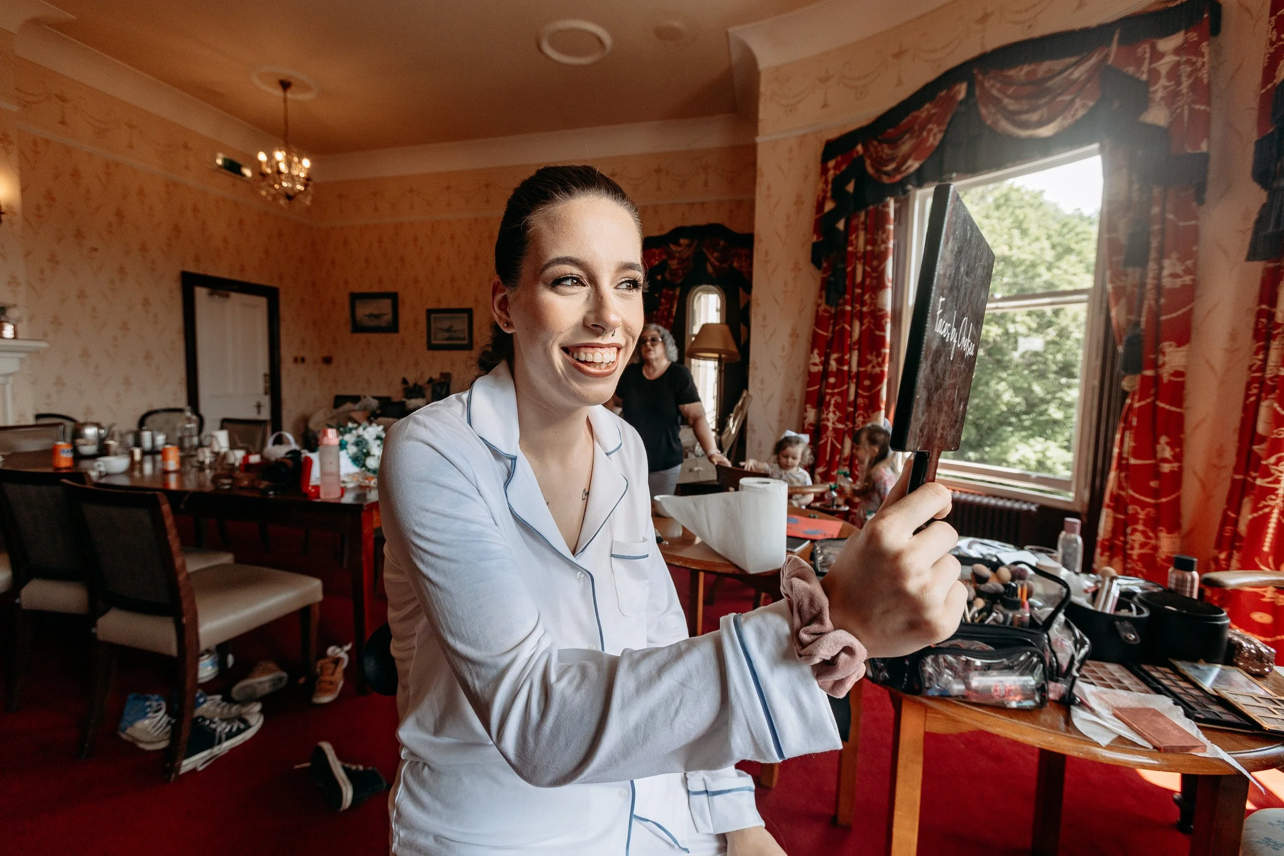 A woman in pajamas is smiling while holding a small blackboard sign in a well-lit room with red curtains, a large window, and children and an older woman in the background.