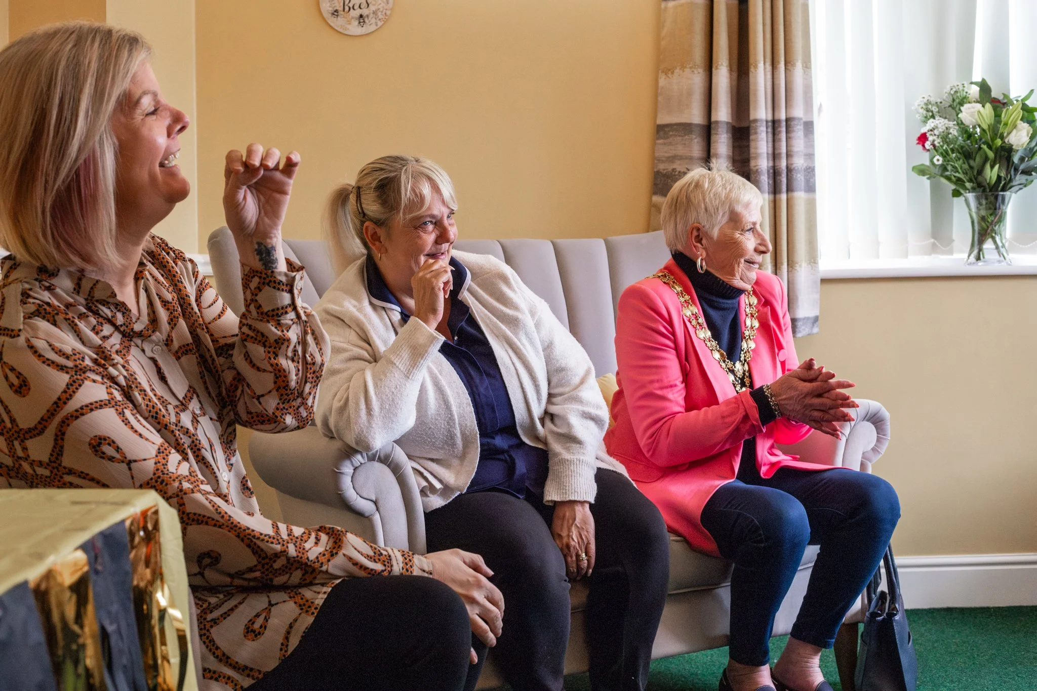 Four women sitting on a sofa in a living room, two are laughing, one looks amused, and the fourth, an older woman with a chain of office, claps her hands, suggesting a celebratory or formal gathering.
