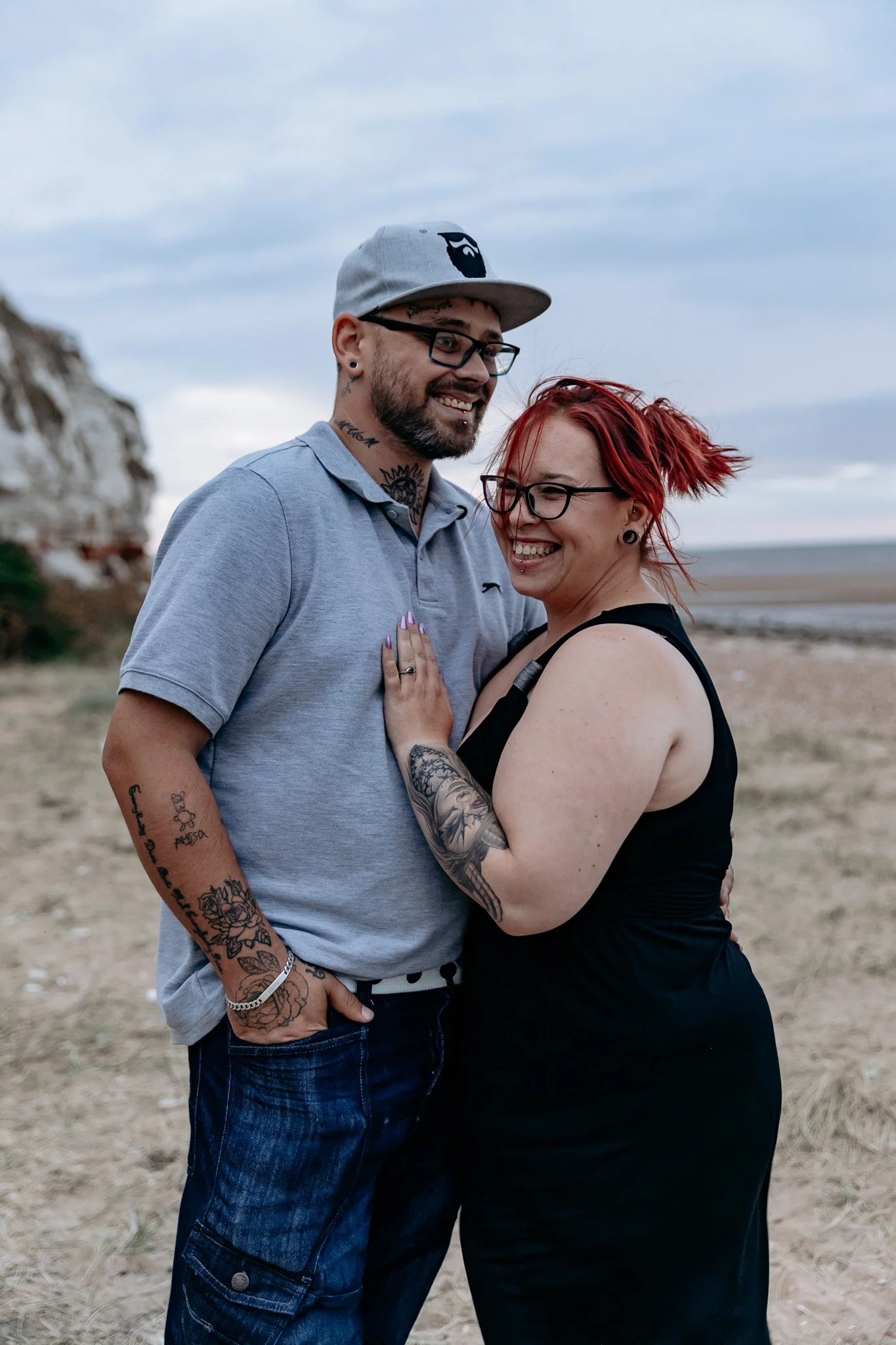 A smiling couple embracing on a beach, with the woman touching the man's chest. The man is wearing a gray polo shirt, jeans, a cap, and glasses, while the woman has red hair, glasses, and tattoos.