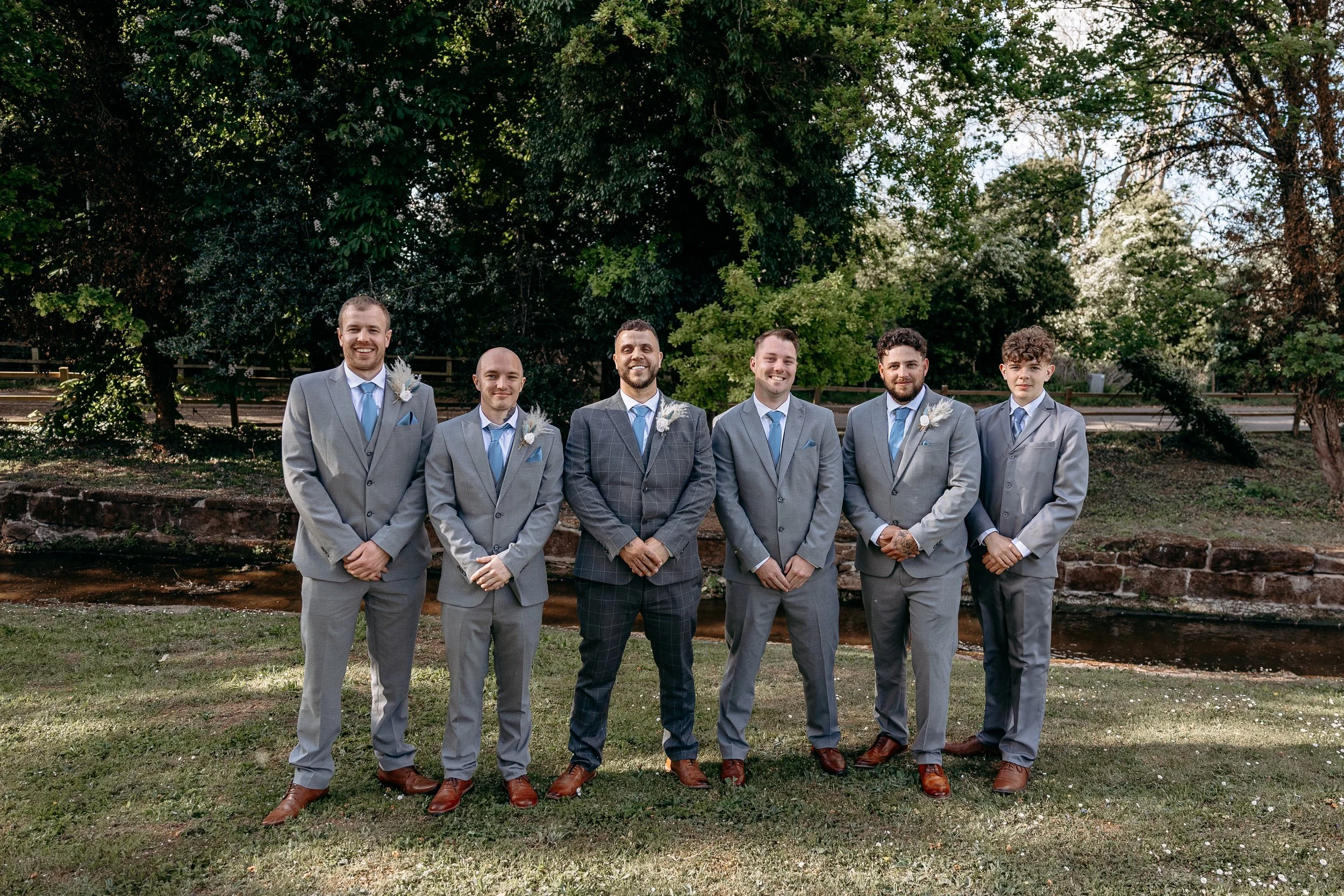 Six men in suits standing outdoors on grass with trees and a small creek in the background.