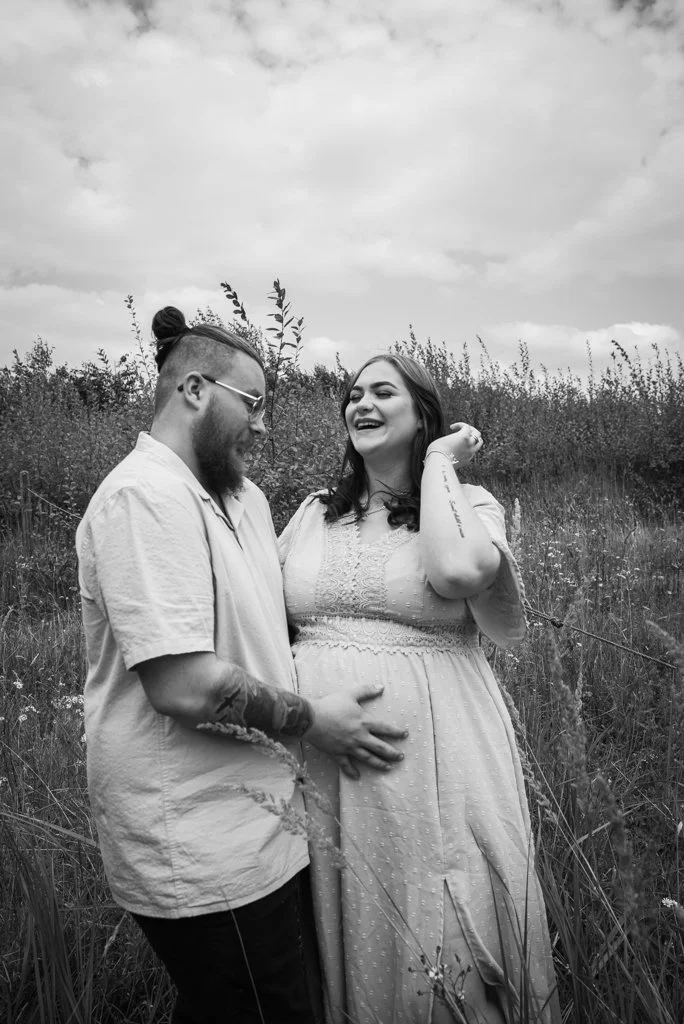 Black and white photo of a pregnant woman and a man in a grassy field. The man is touching the woman's belly, and they are both smiling, sharing a joyful moment.