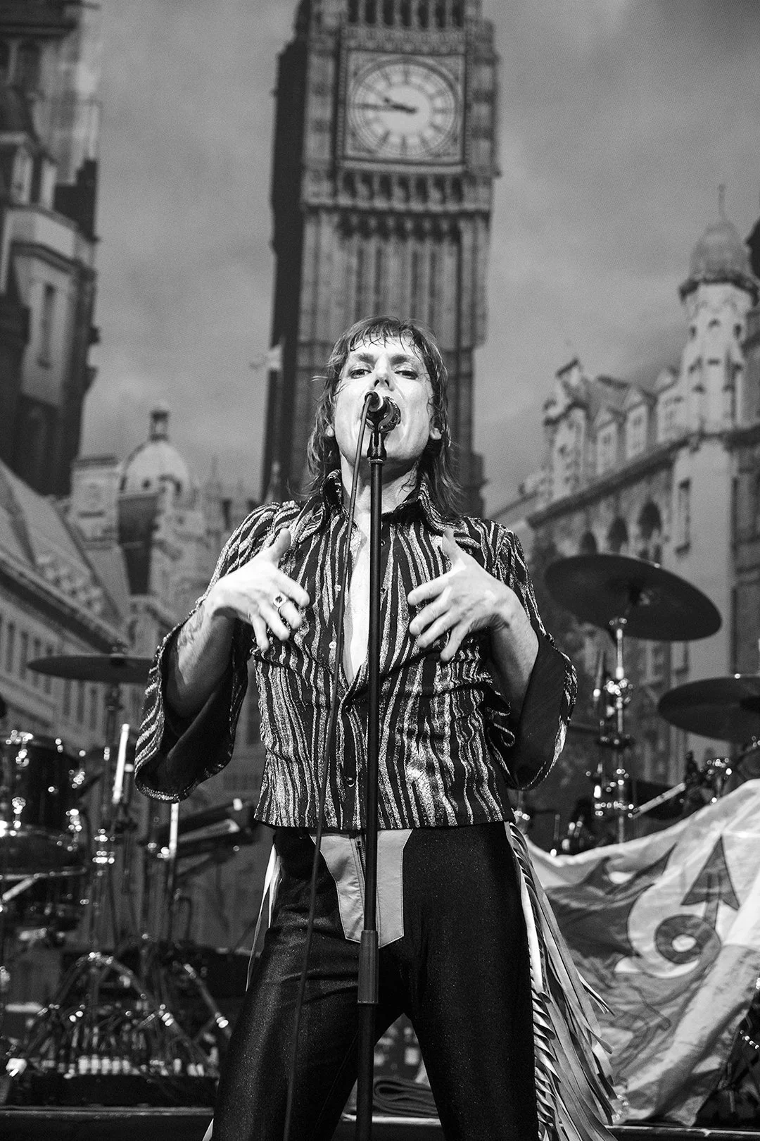 Black and white photo of a performer singing on stage, wearing a striped outfit, with a backdrop of Big Ben and London architecture.