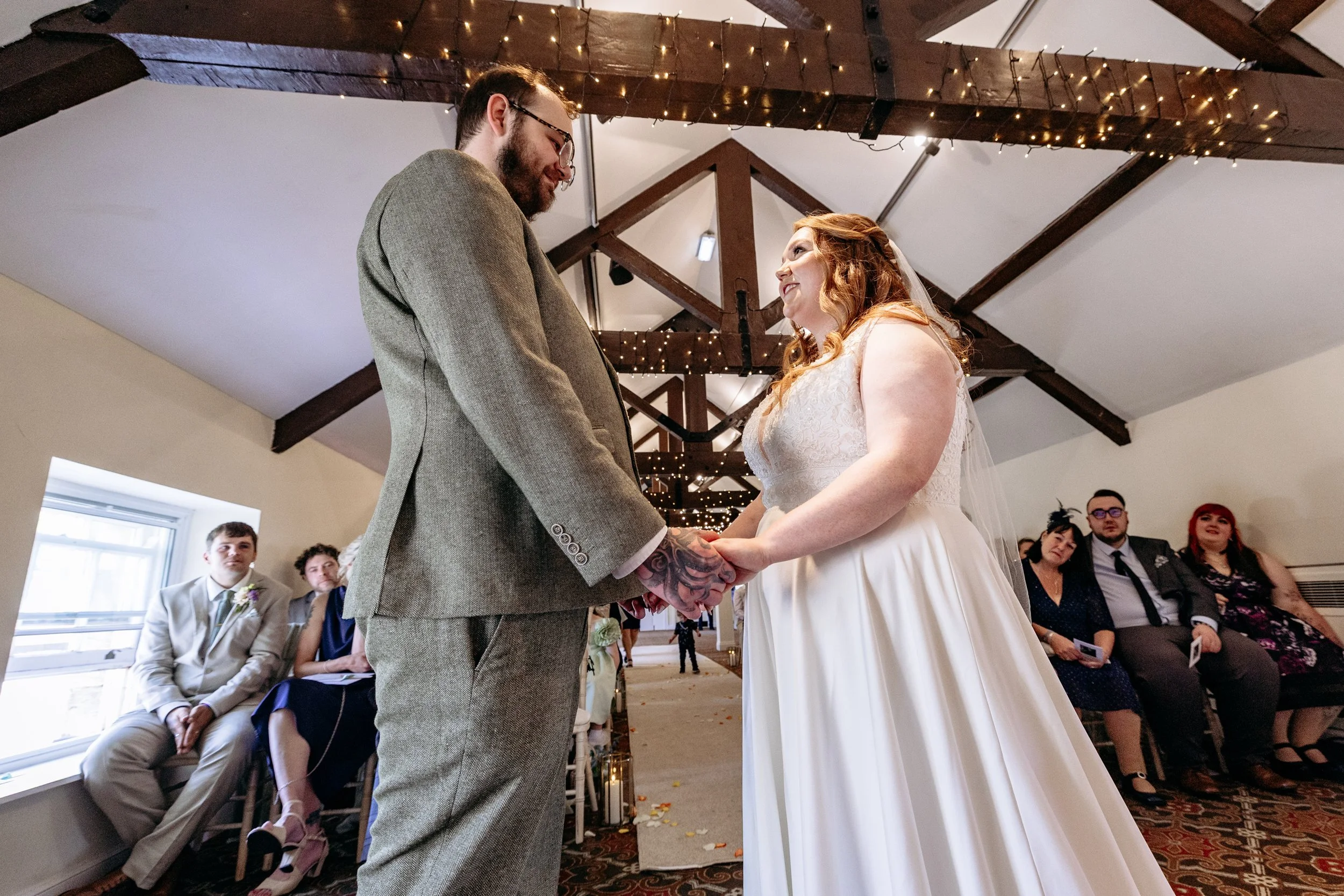 A bride and groom holding hands and gazing at each other during their wedding ceremony, with seated guests in the background, inside a rustic venue decorated with string lights.