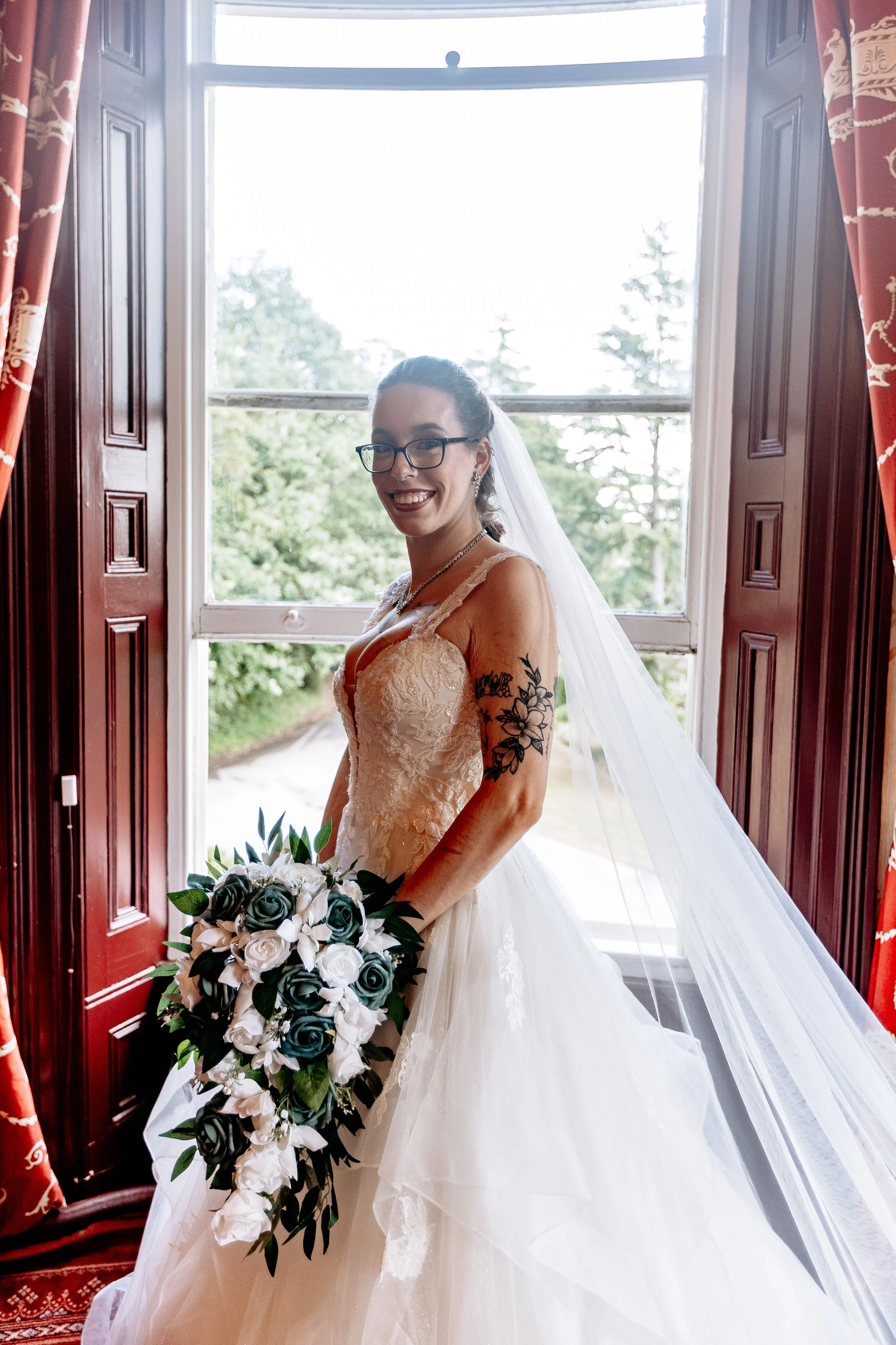A bride standing by a window, smiling, wearing glasses, a lace wedding dress, and holding a bouquet of white and dark-colored flowers.