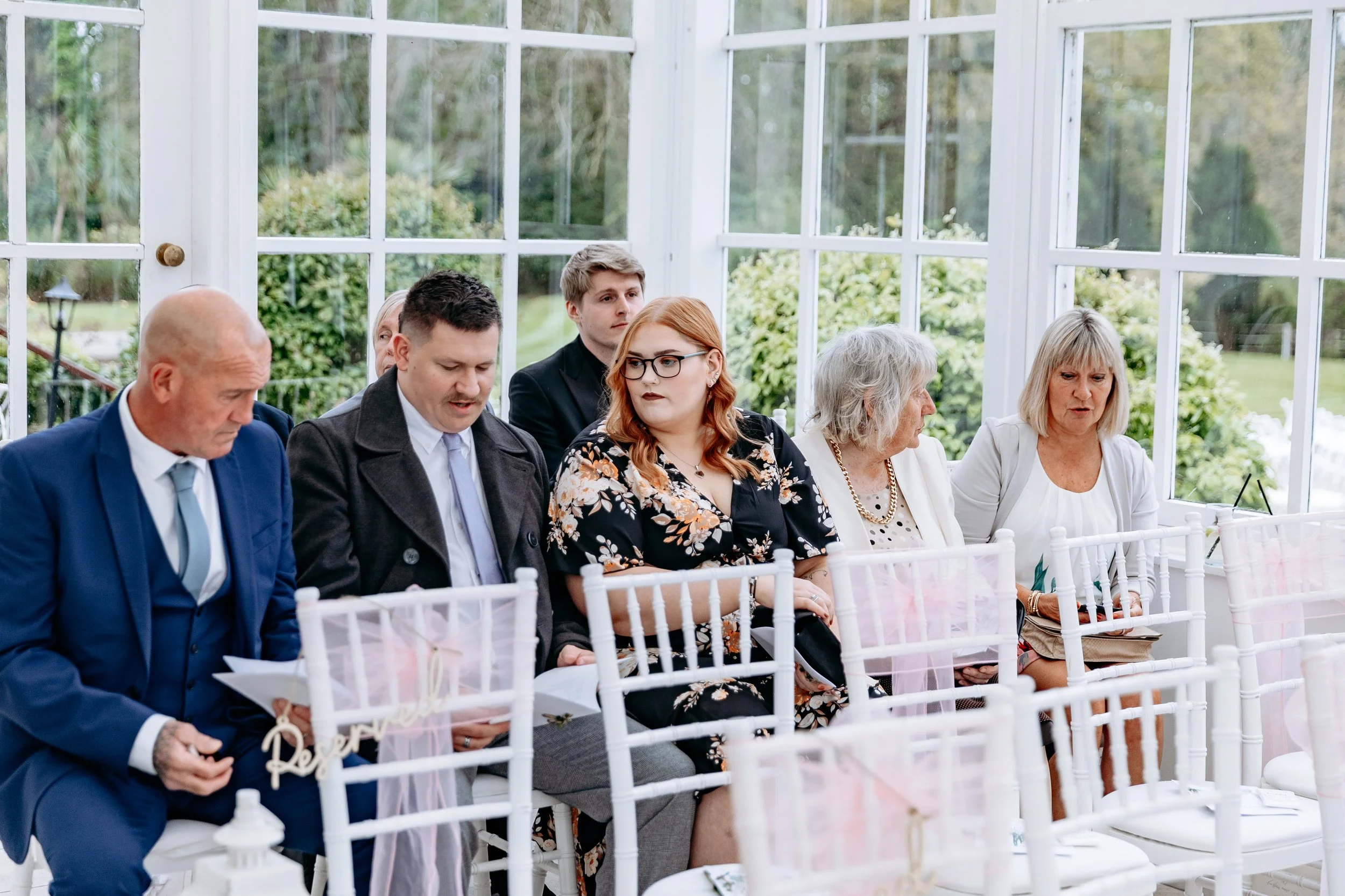 Group of people sitting in a conservatory with glass walls, attending a formal event, most are reading or looking at programs.