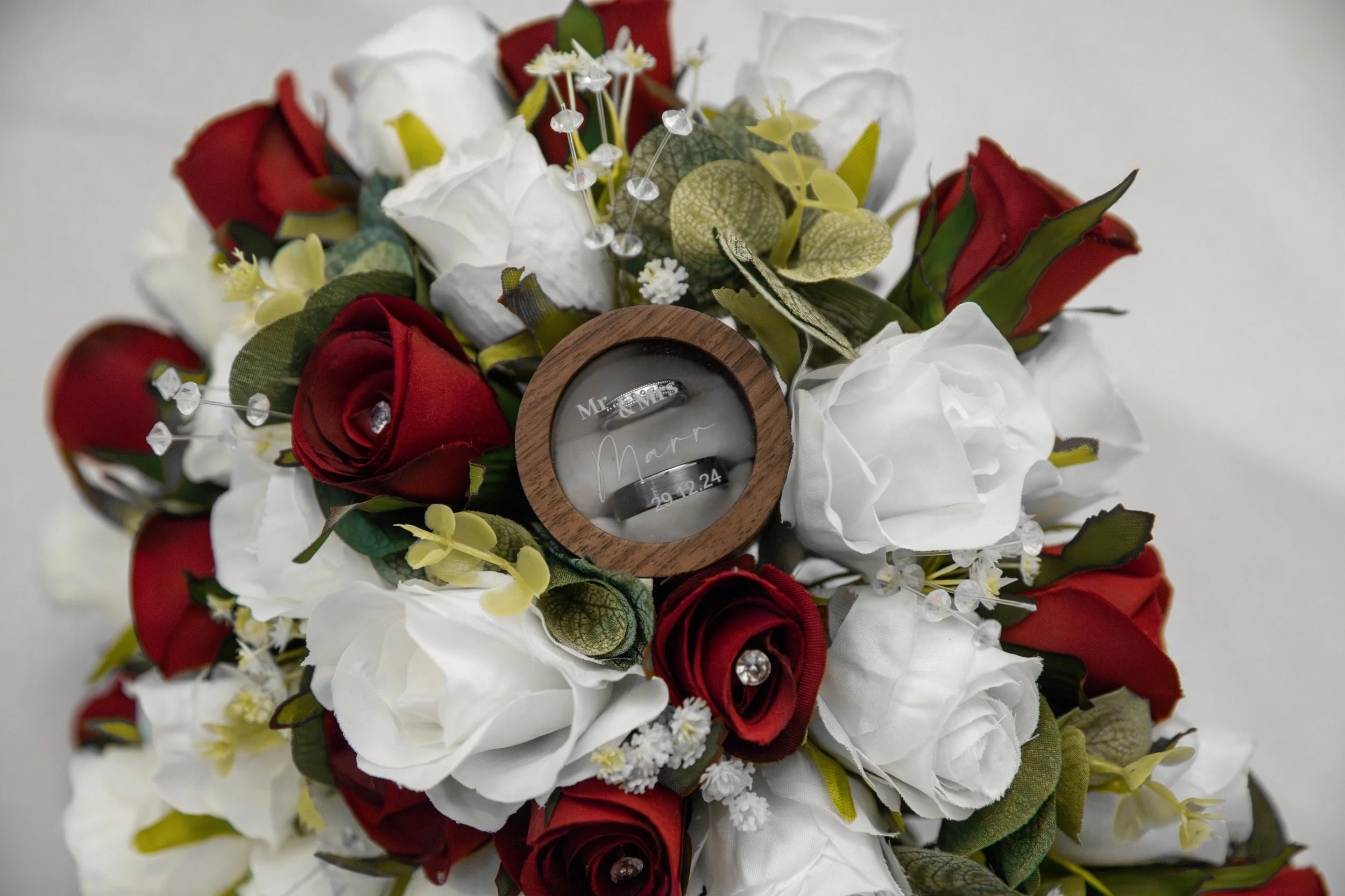 A bridal bouquet with red and white roses, green foliage, and decorative elements, including a round wooden frame with an engraved message and a watch inside.