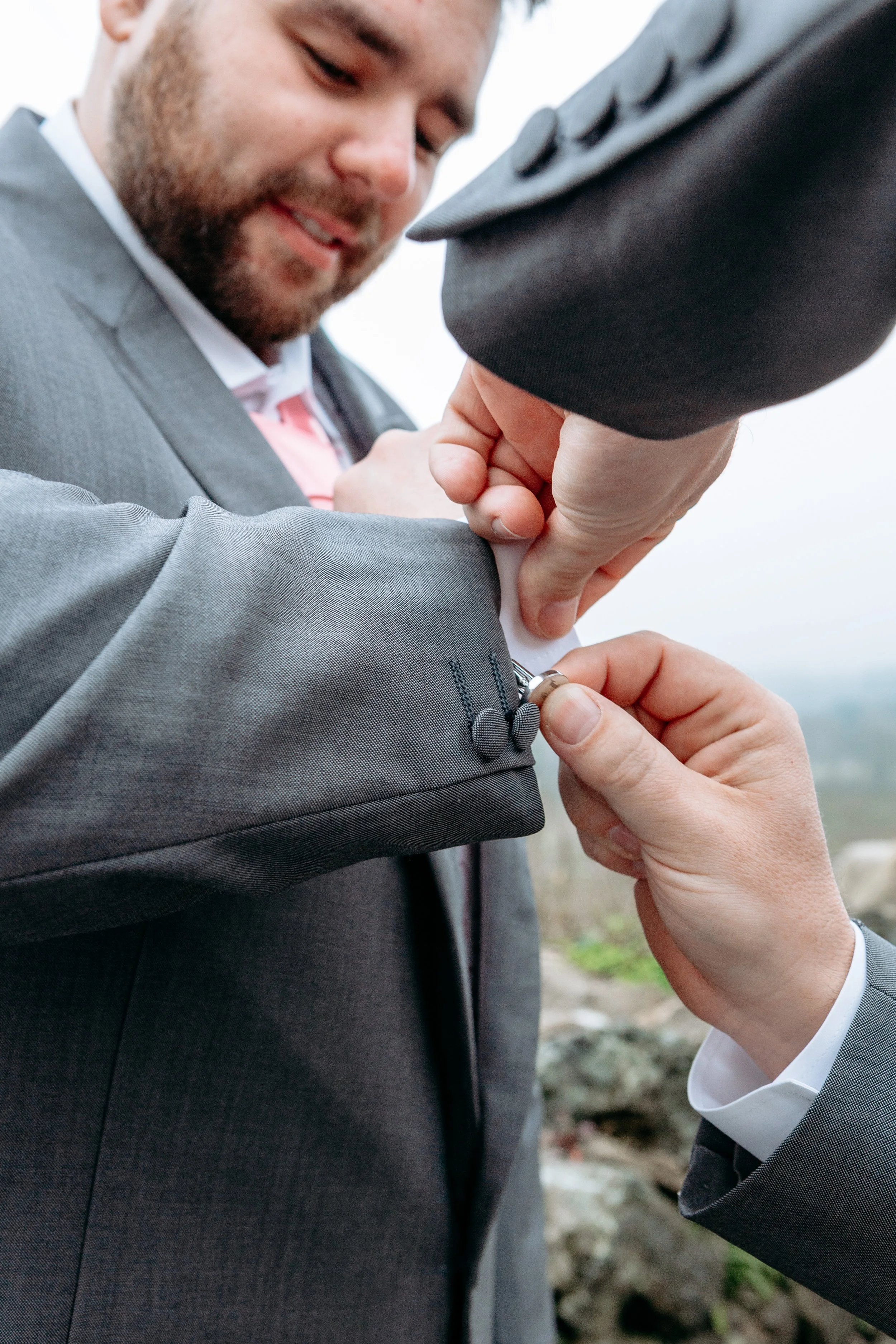 Person in a gray suit getting cufflinks fastened by another person in a suit, smiling.