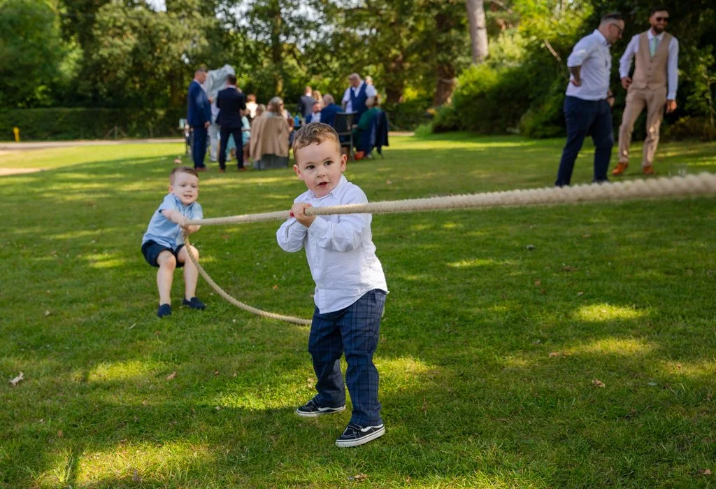 Two young boys are playing tug-of-war with a rope on a grassy lawn during an outdoor celebration, with adults gathered in the background.