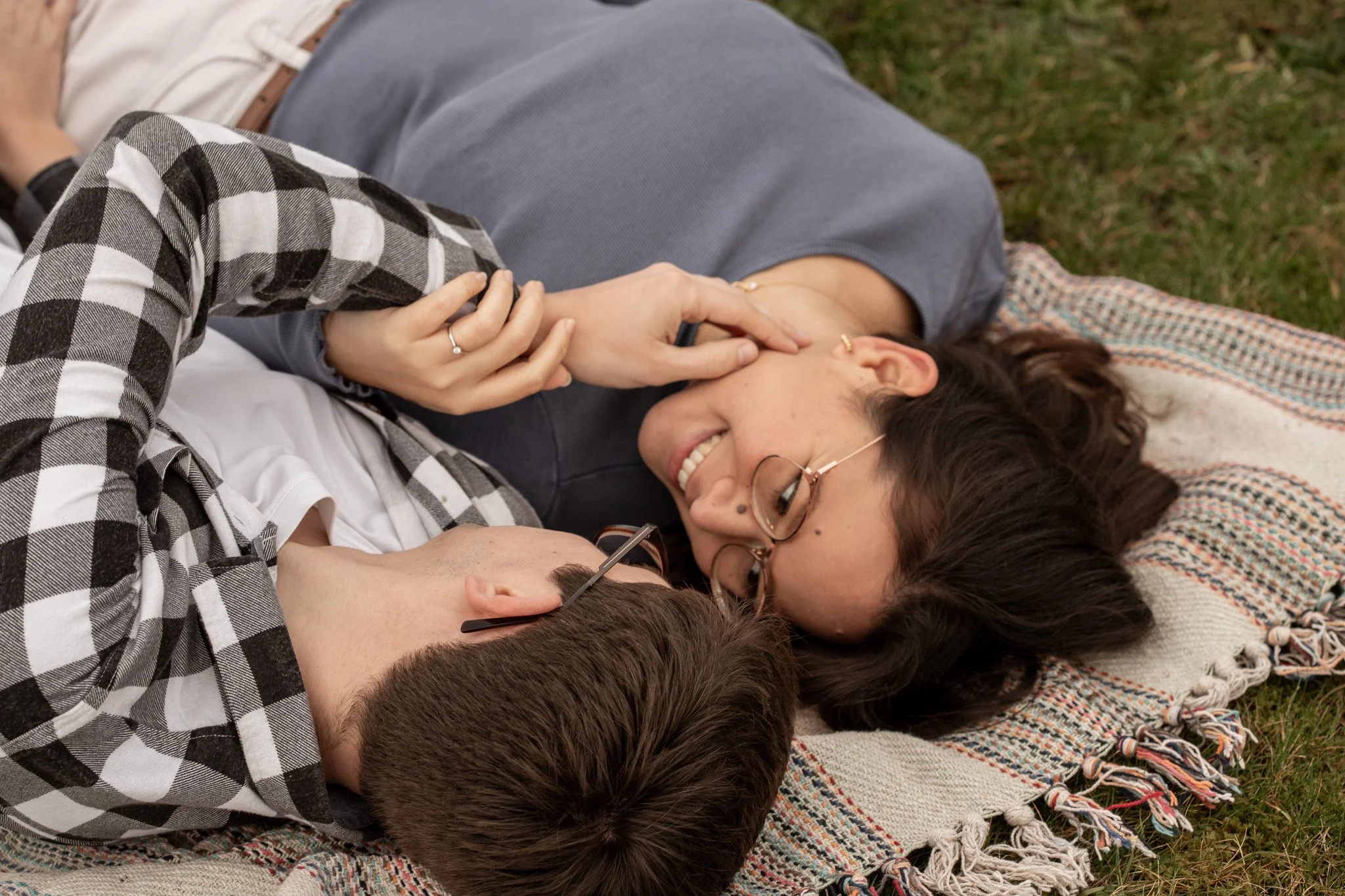 A young couple lying on a blanket outside, smiling and looking at each other, with the woman gently touching the man's face.