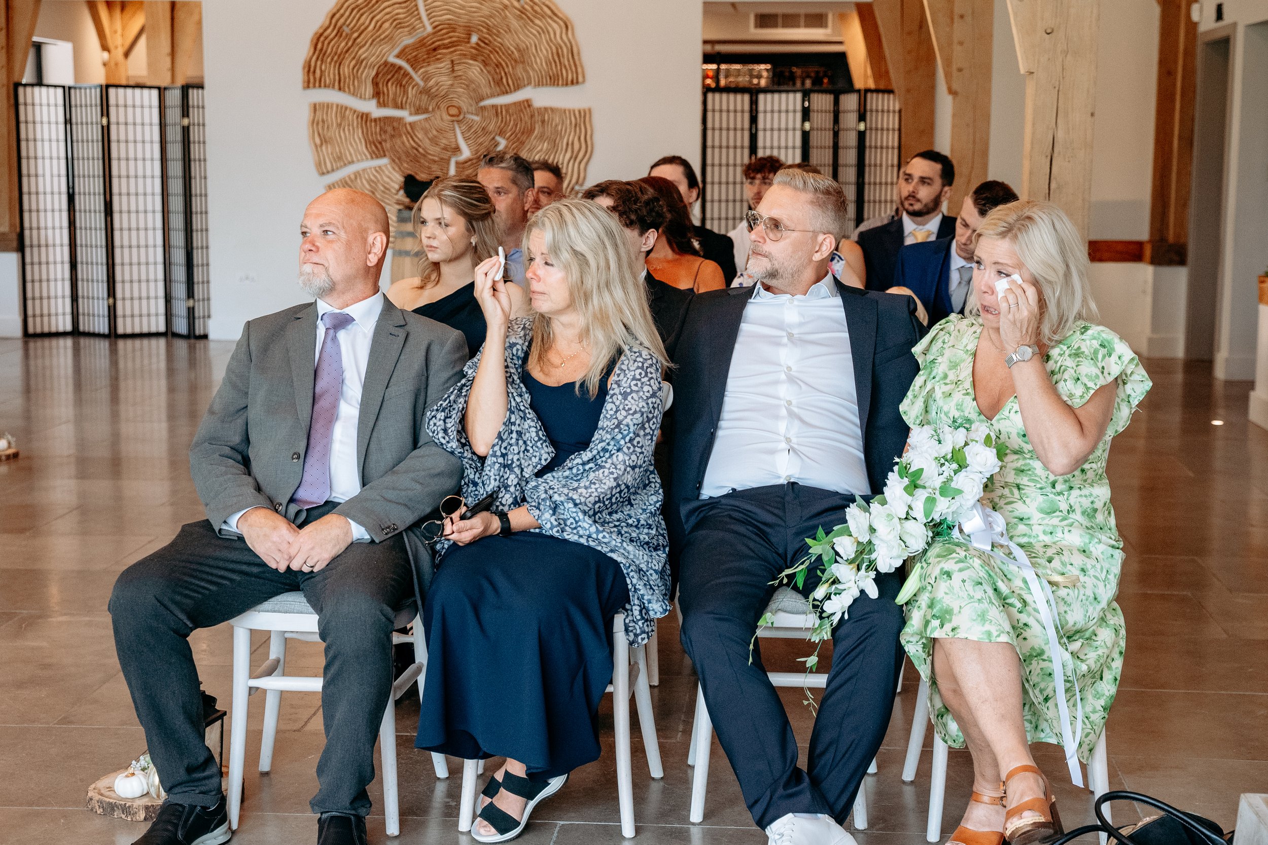 A group of people sitting in a brightly lit room during an emotional event, with some wiping away tears. The setting includes a decorative wooden wall piece in the background.
