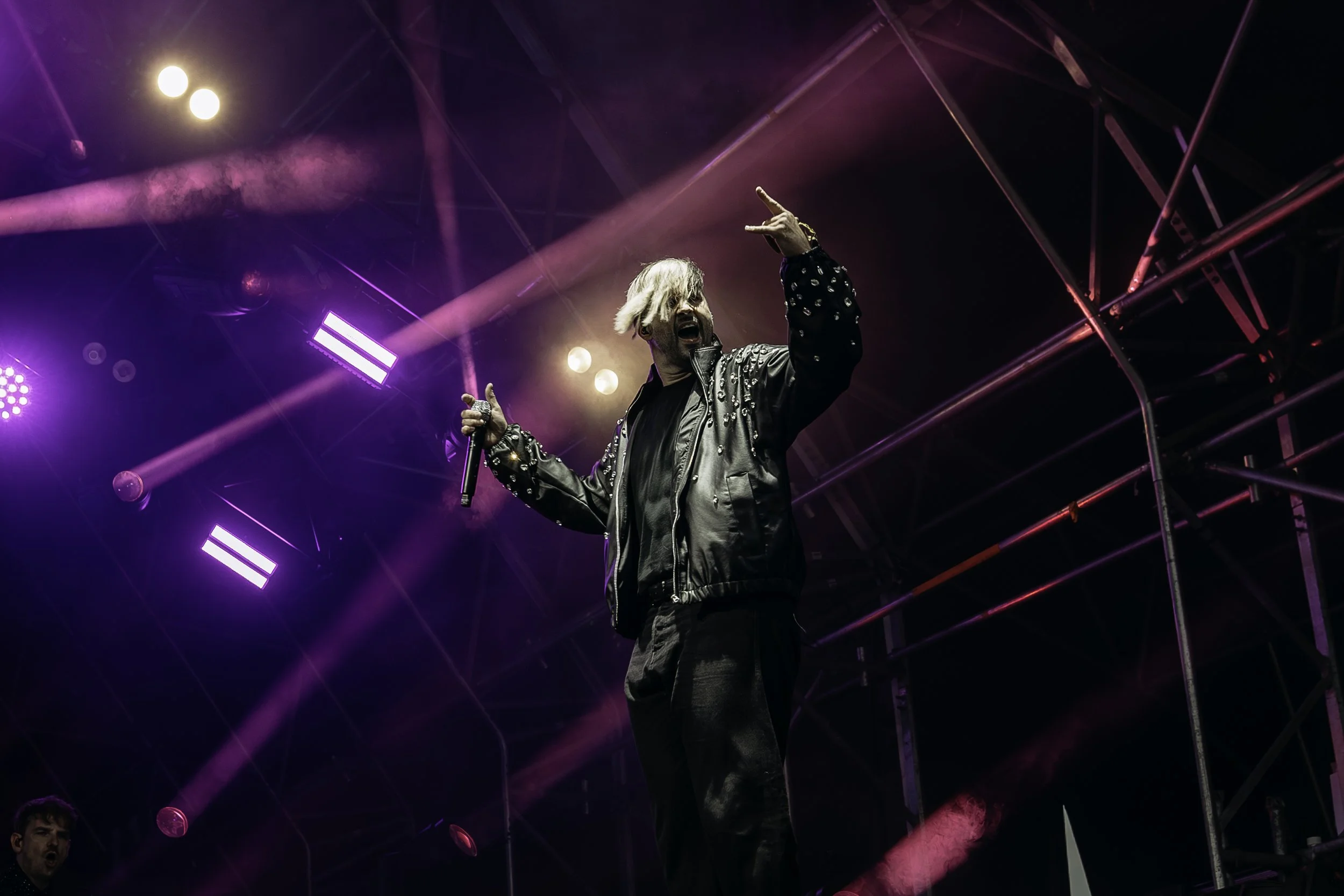 Performer with blonde hair singing on stage with purple and pink lighting and scaffolding in the background.