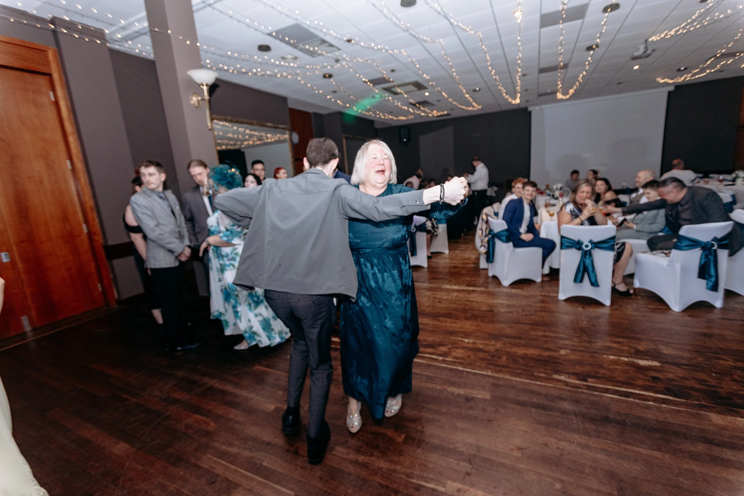 People dancing and socializing at a formal event or celebration in a decorated banquet hall with string lights and tables.