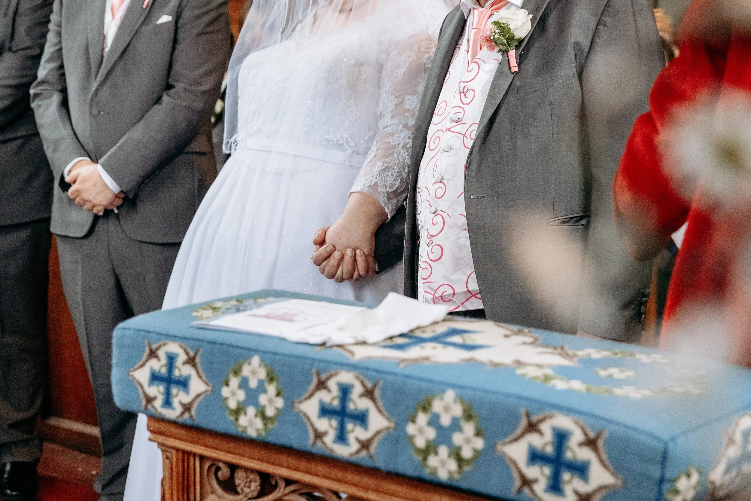 A bride and groom holding hands during a wedding ceremony with a priest officiating, with a decorated table in the foreground.