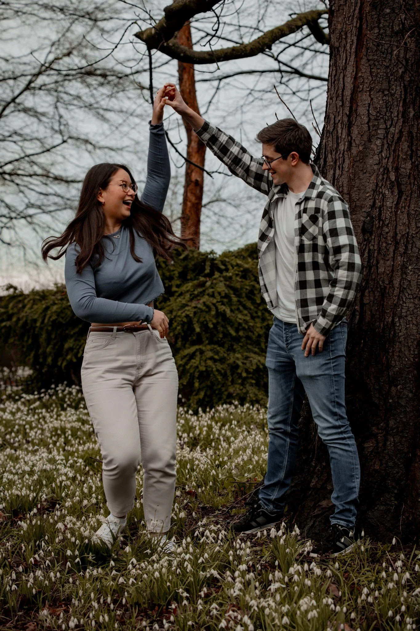 A man and woman are dancing and smiling outdoors near a large tree and blooming snowdrop flowers, enjoying each other's company.