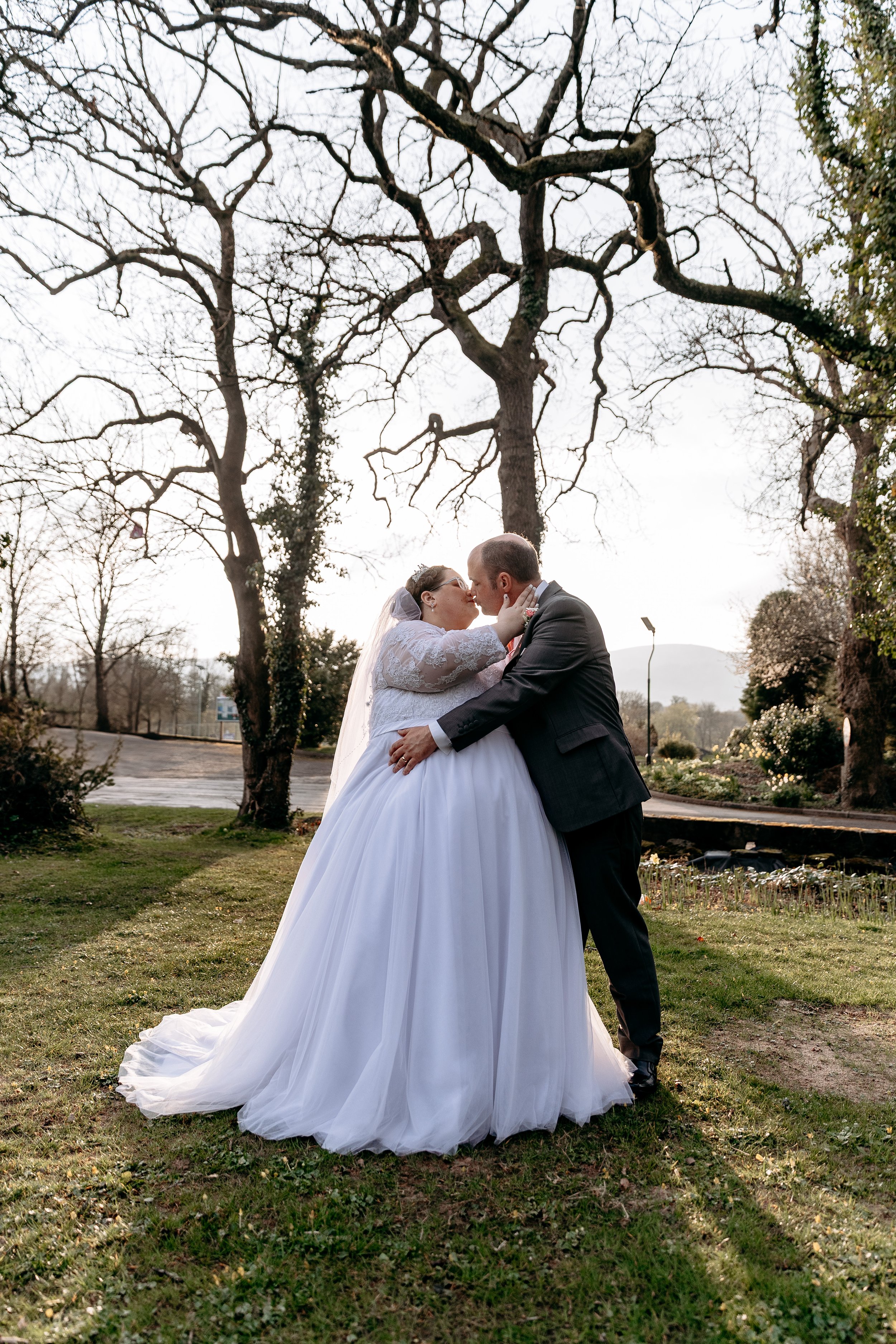 A bride and groom sharing a kiss outdoors on their wedding day, with leafless trees and a cloudy sky in the background.