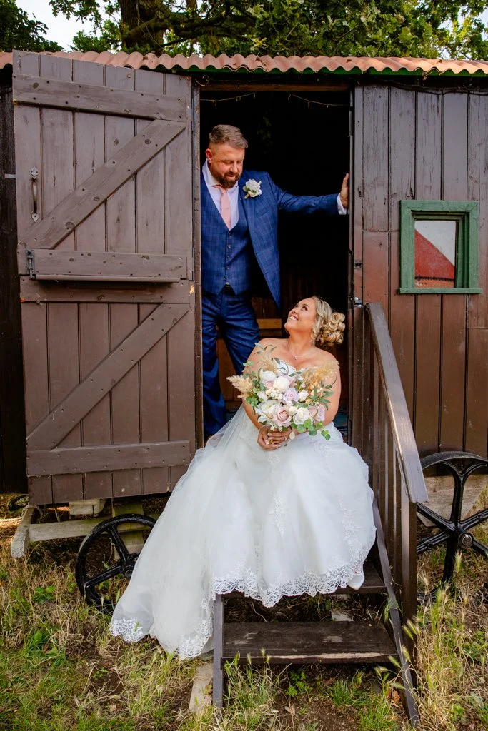 A bride sitting on the steps of a small wooden shed, holding a bouquet, looking up at a groom standing at the doorway, dressed in a blue suit with a boutonniere, smiling at her.
