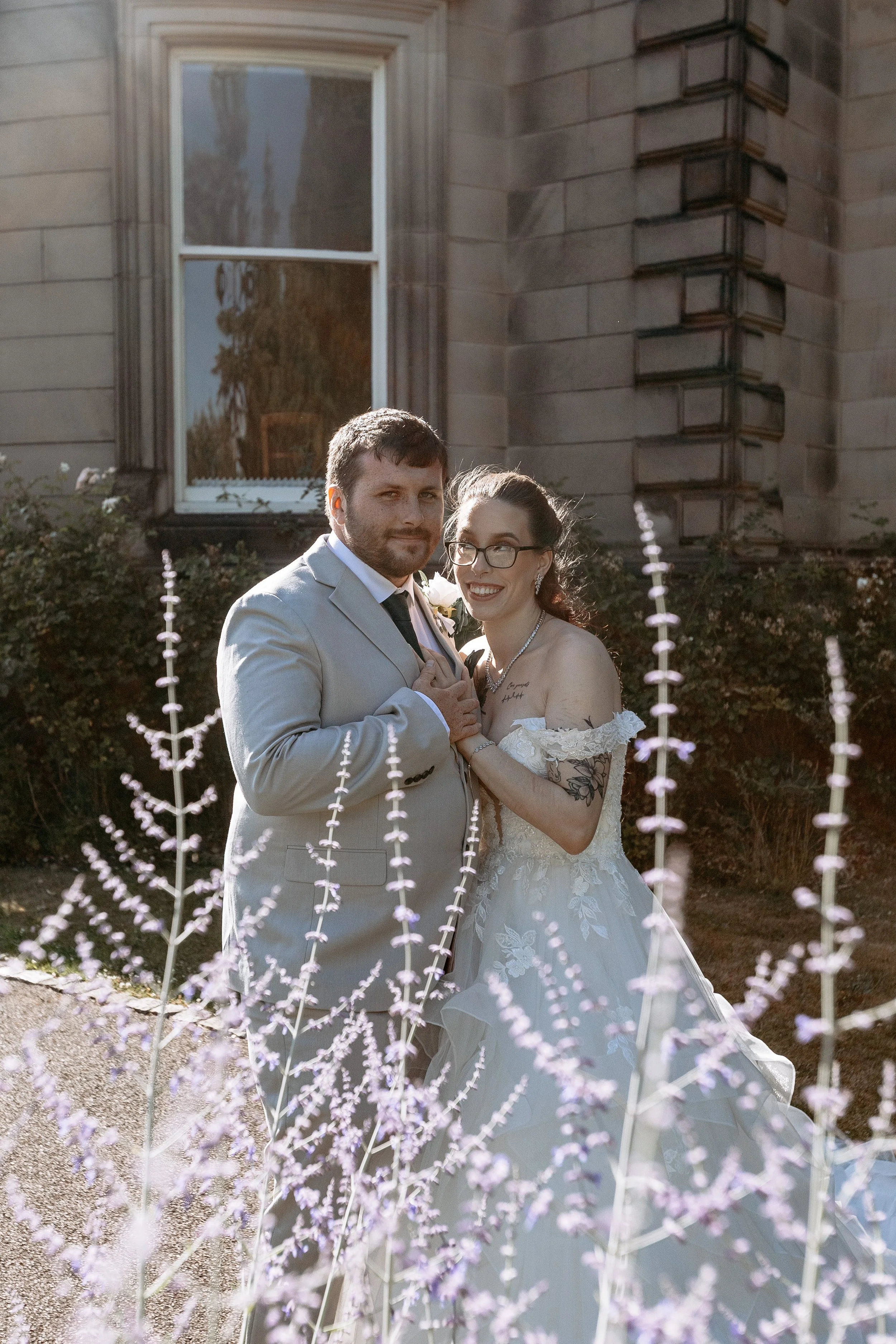 A newlywed couple stands together outdoors in front of an old stone building with a window, surrounded by purple flowers. The bride is wearing glasses and a white lace wedding dress, and the groom is in a light gray suit with a white shirt and dark t