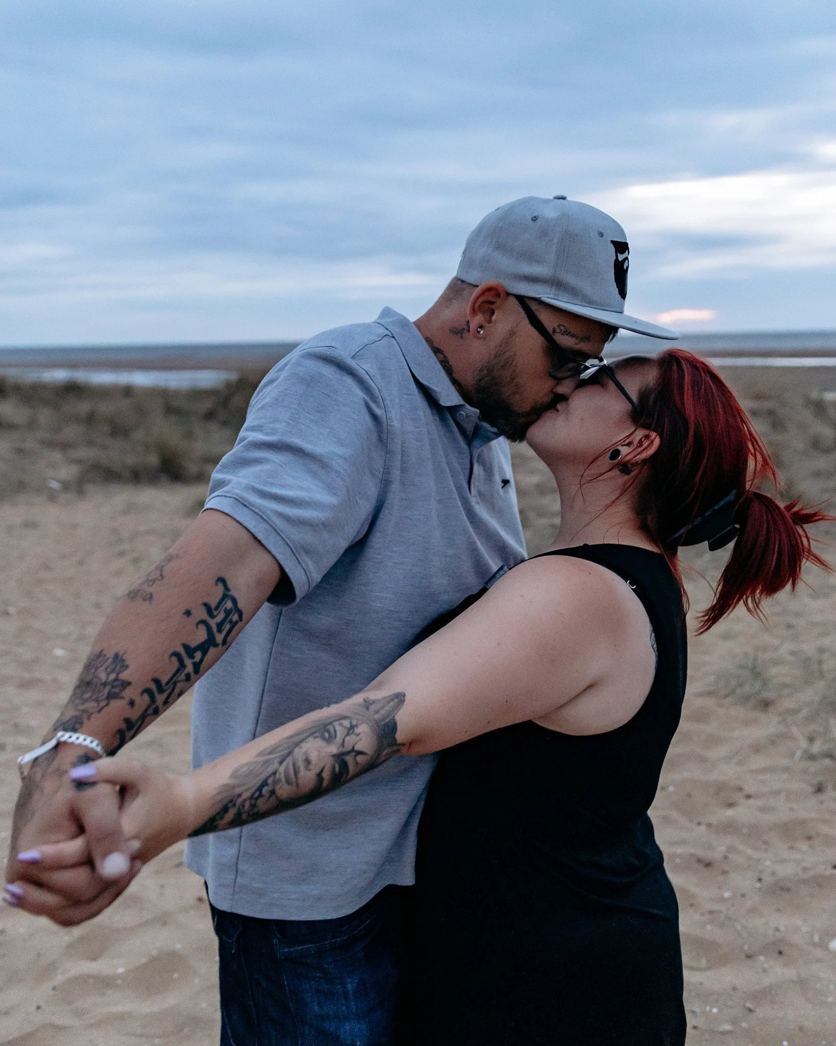 A man and woman sharing a kiss on a beach at sunset, holding hands and wearing casual clothing and sunglasses.