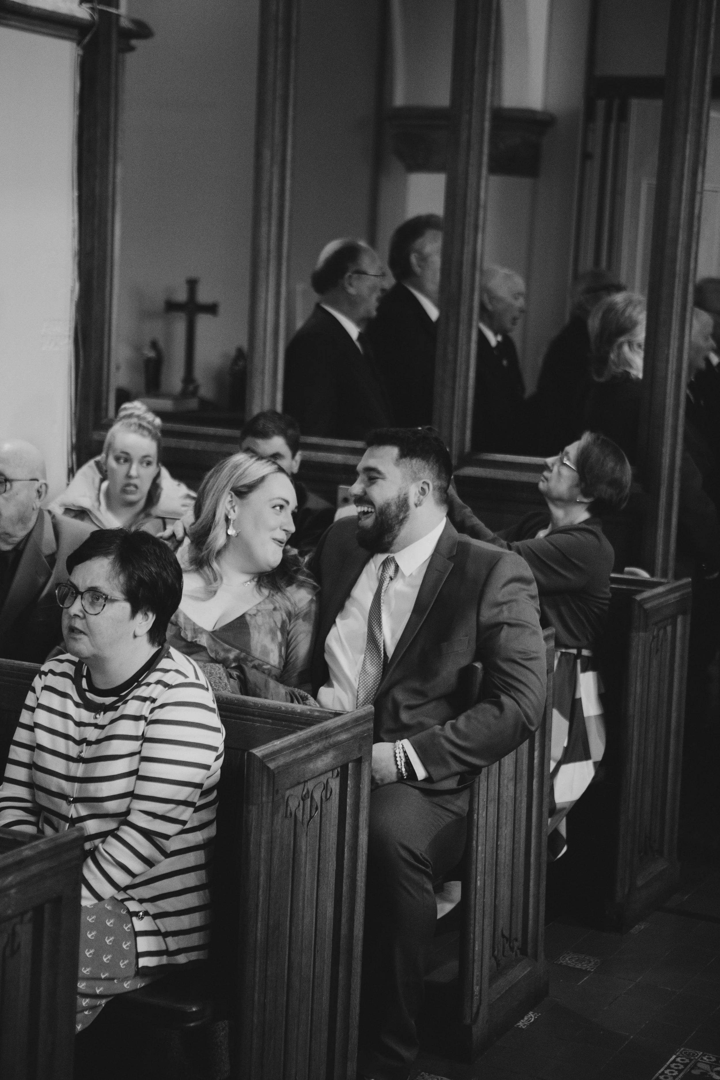 People sitting in pews inside a church, some are smiling, and others are engaged in conversation, with a reflection of six men standing and praying in a glass partition behind them.