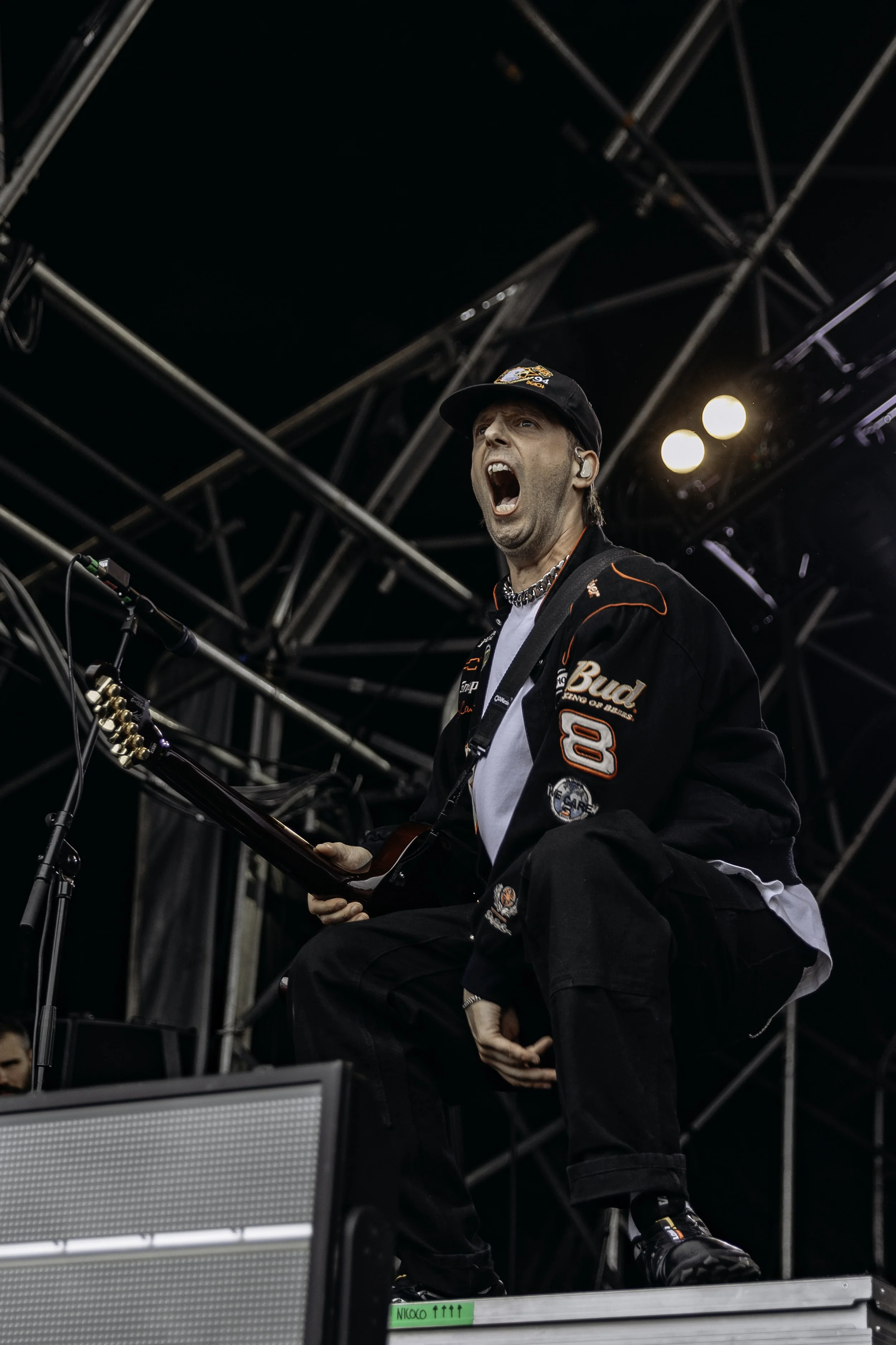 A male musician on stage, singing passionately with an open mouth, wearing a black baseball cap, a black jacket with patches, sunglasses, and holding a guitar.