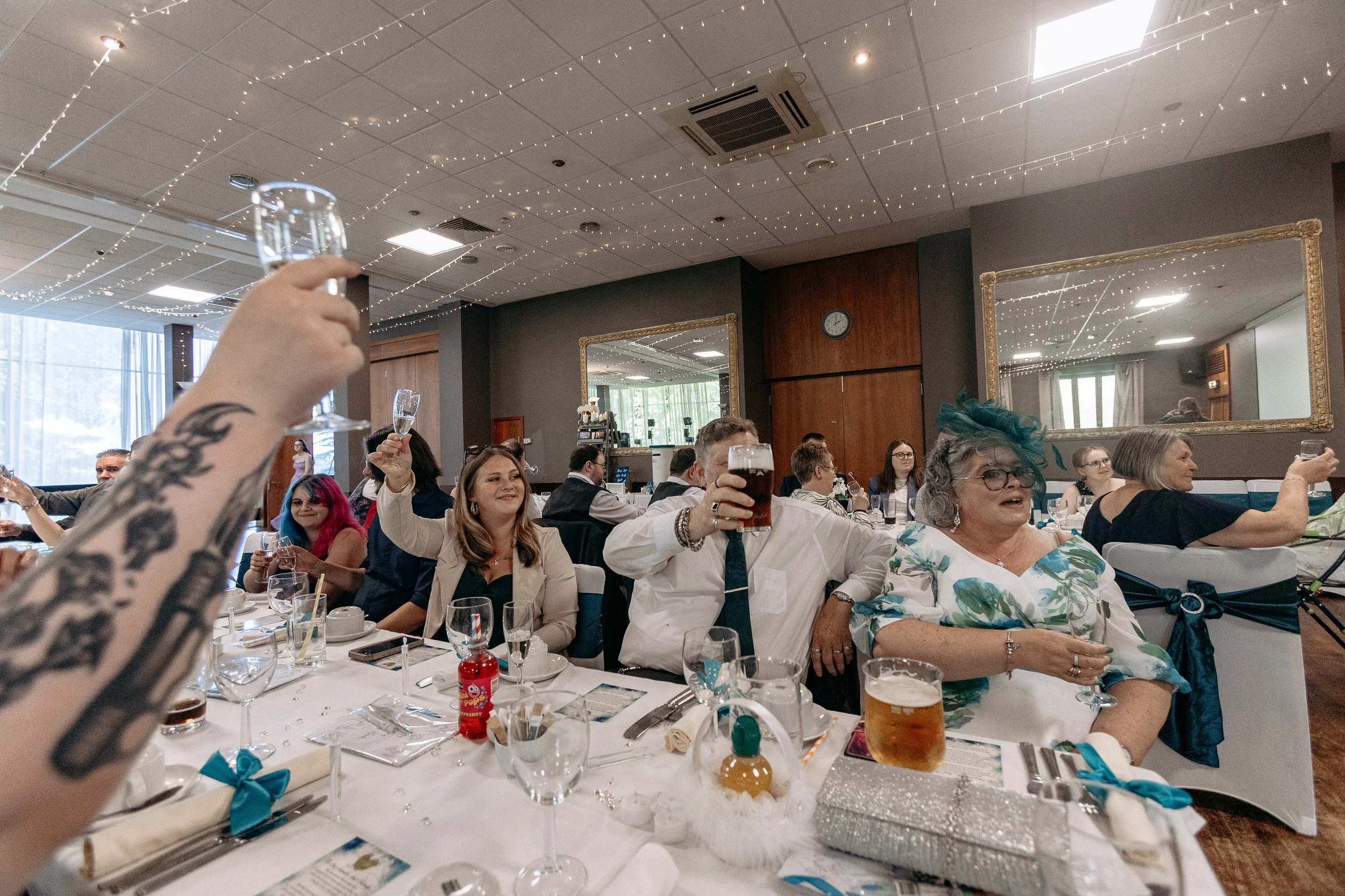 People celebrating at a banquet table, toasting with drinks, in a decorated event hall with fairy lights and mirrors.