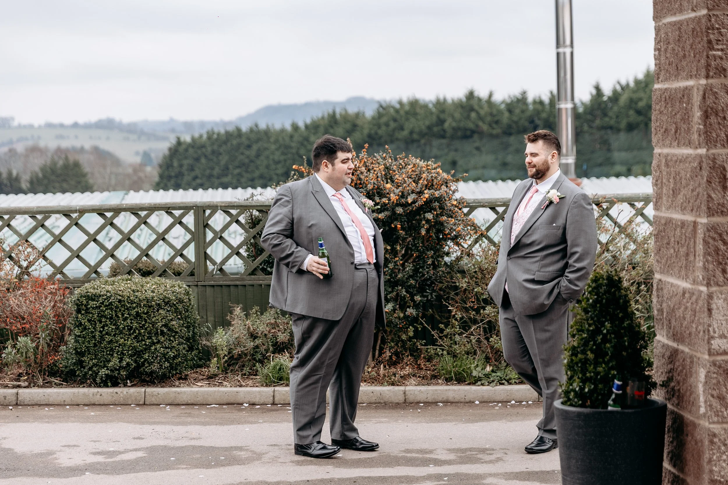 Two men in gray suits standing outside near a brick wall and a garden, engaged in conversation at a wedding or formal event, with a scenic landscape in the background.