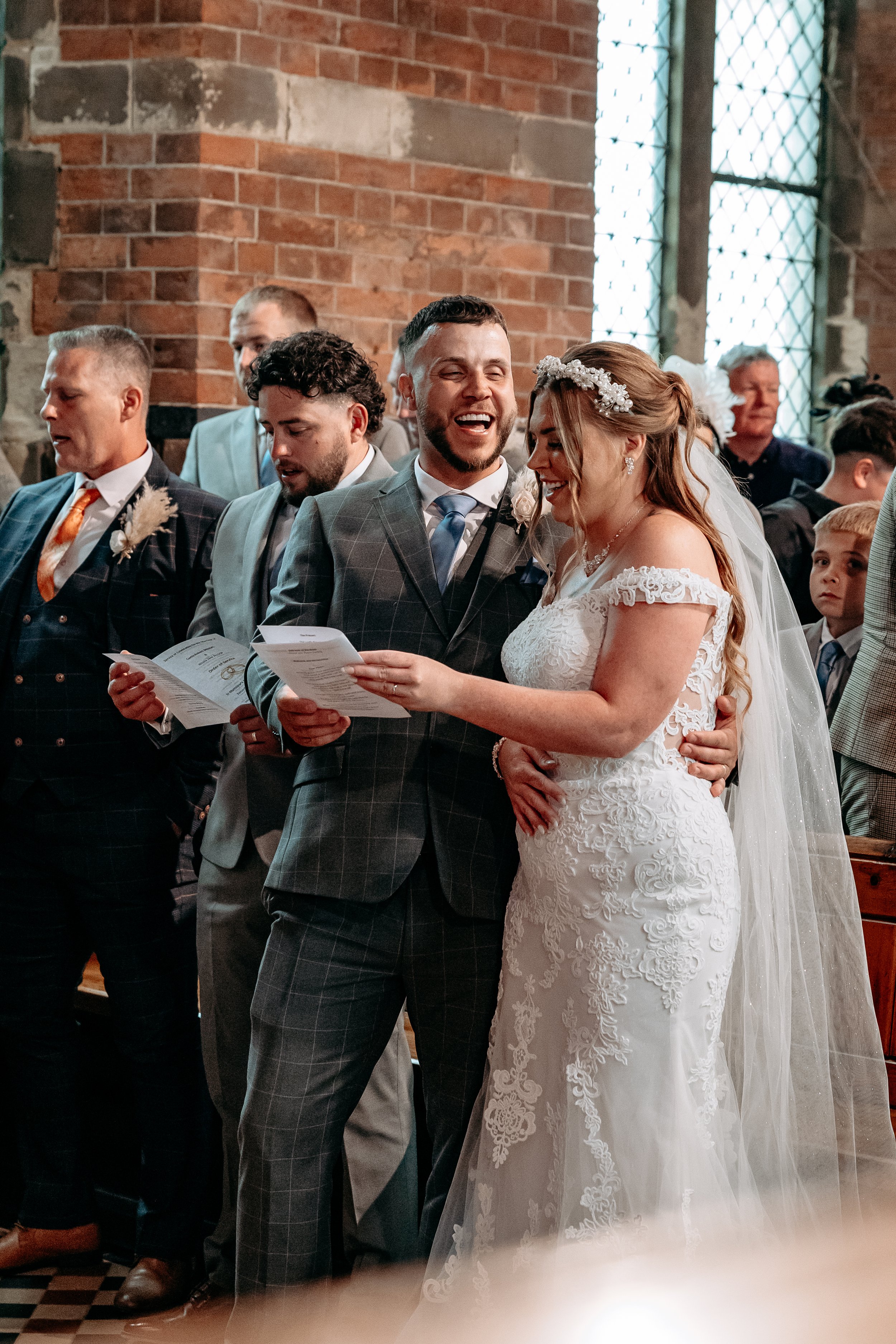 A bride and groom sharing a joyful moment during their wedding ceremony inside a church with brick walls and stained glass windows, surrounded by guests.