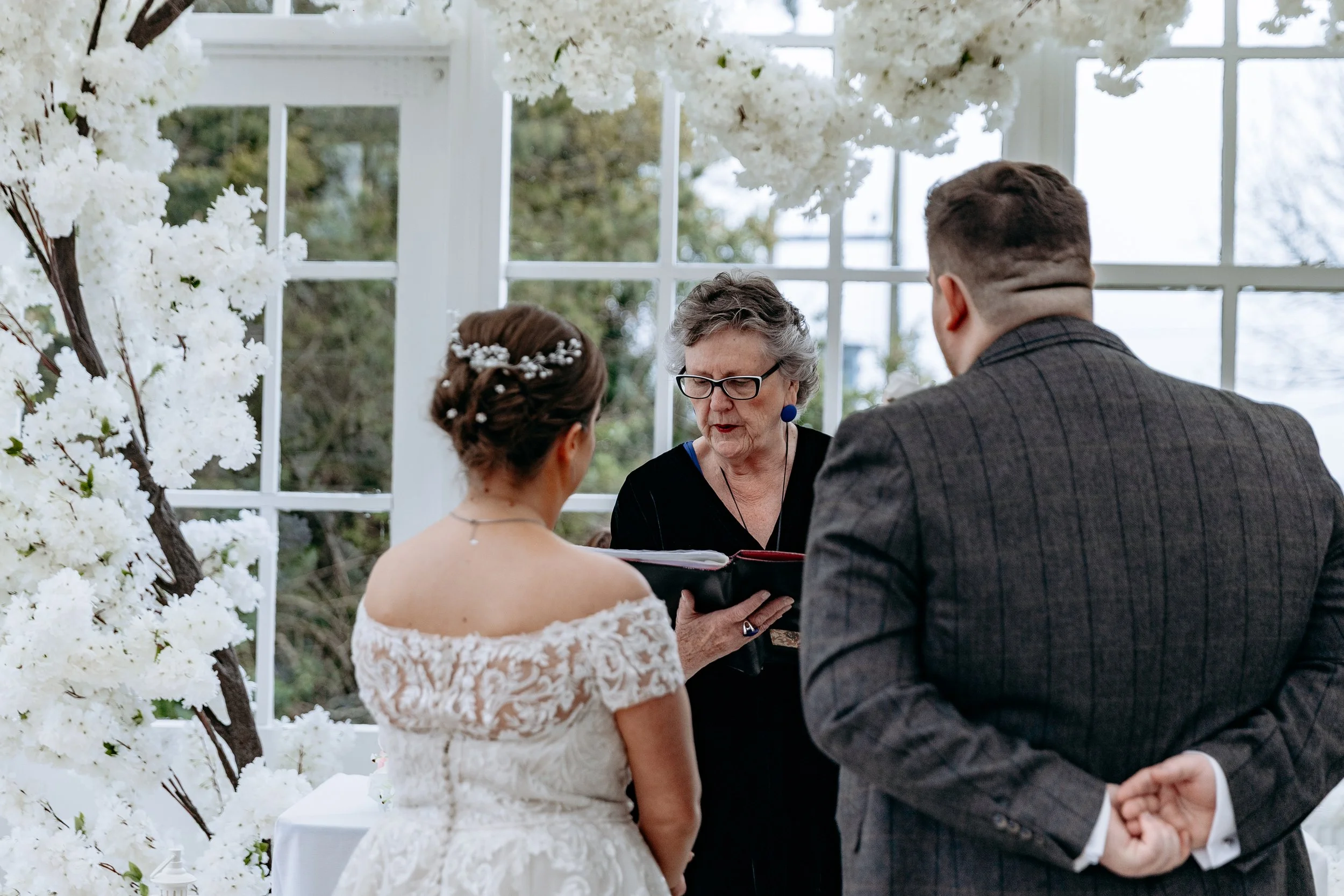 A wedding ceremony with a bride and groom standing before an officiant in a glass-enclosed venue decorated with white flowers.