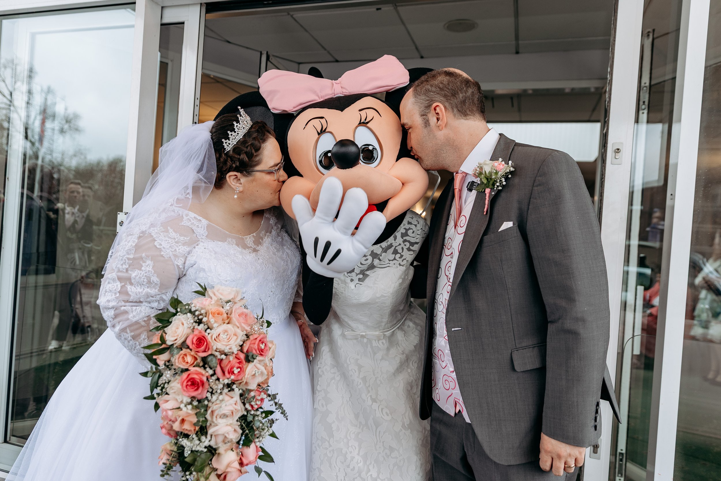 A bride and groom with a woman dressed as Minnie Mouse, all kissing the character's face during a wedding celebration.