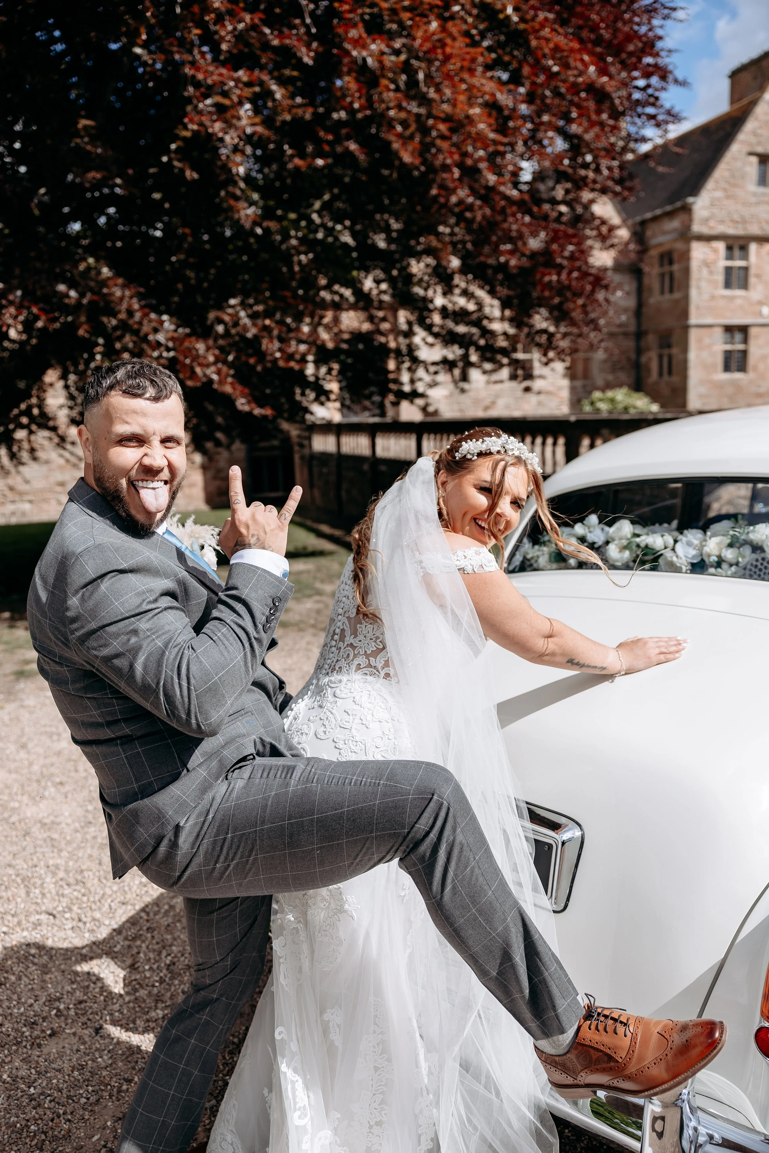 A smiling bride and groom at their wedding, standing next to a vintage white car decorated with flowers in an outdoor setting with trees and historic buildings.