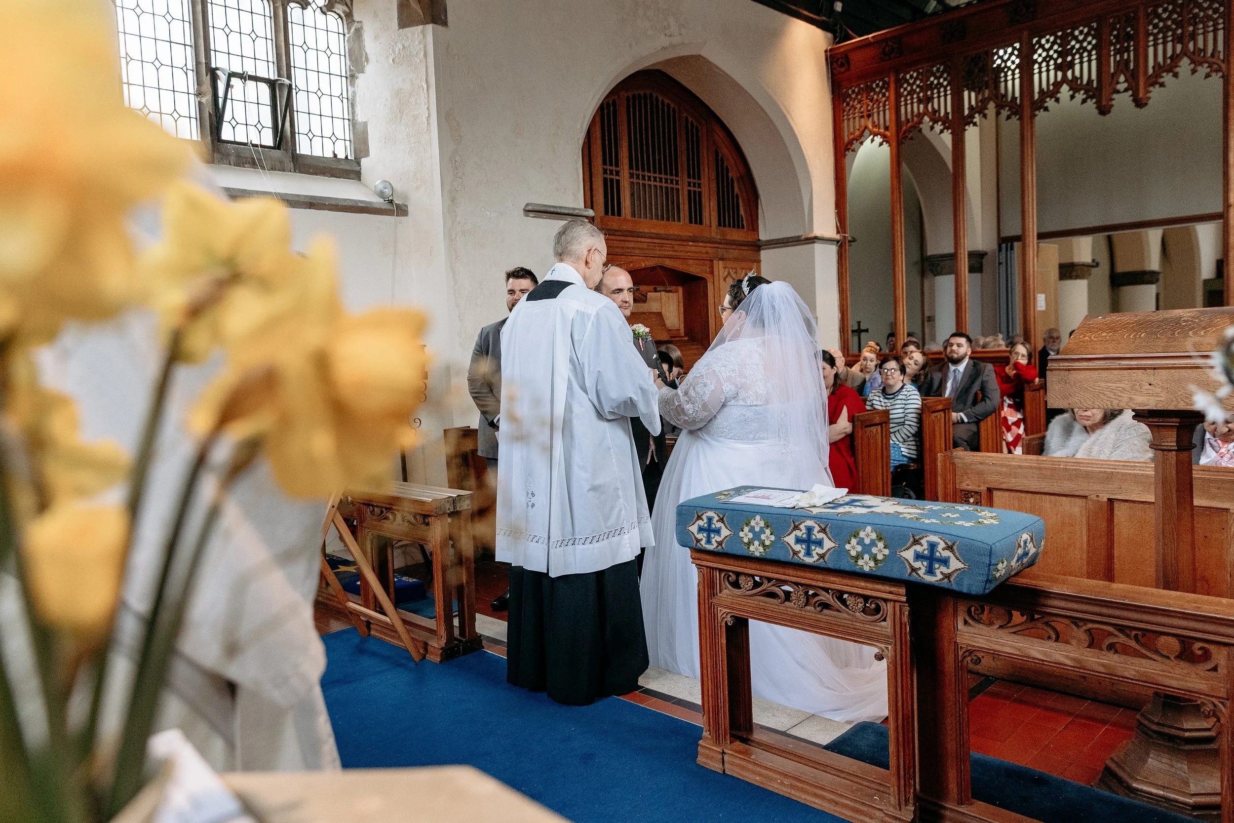 A wedding ceremony inside a church with the bride and groom standing before a priest. The bride wears a white wedding gown and veil, and the groom wears a suit. Guests are seated in wooden pews, watching the ceremony. A blurry bunch of yellow flowers