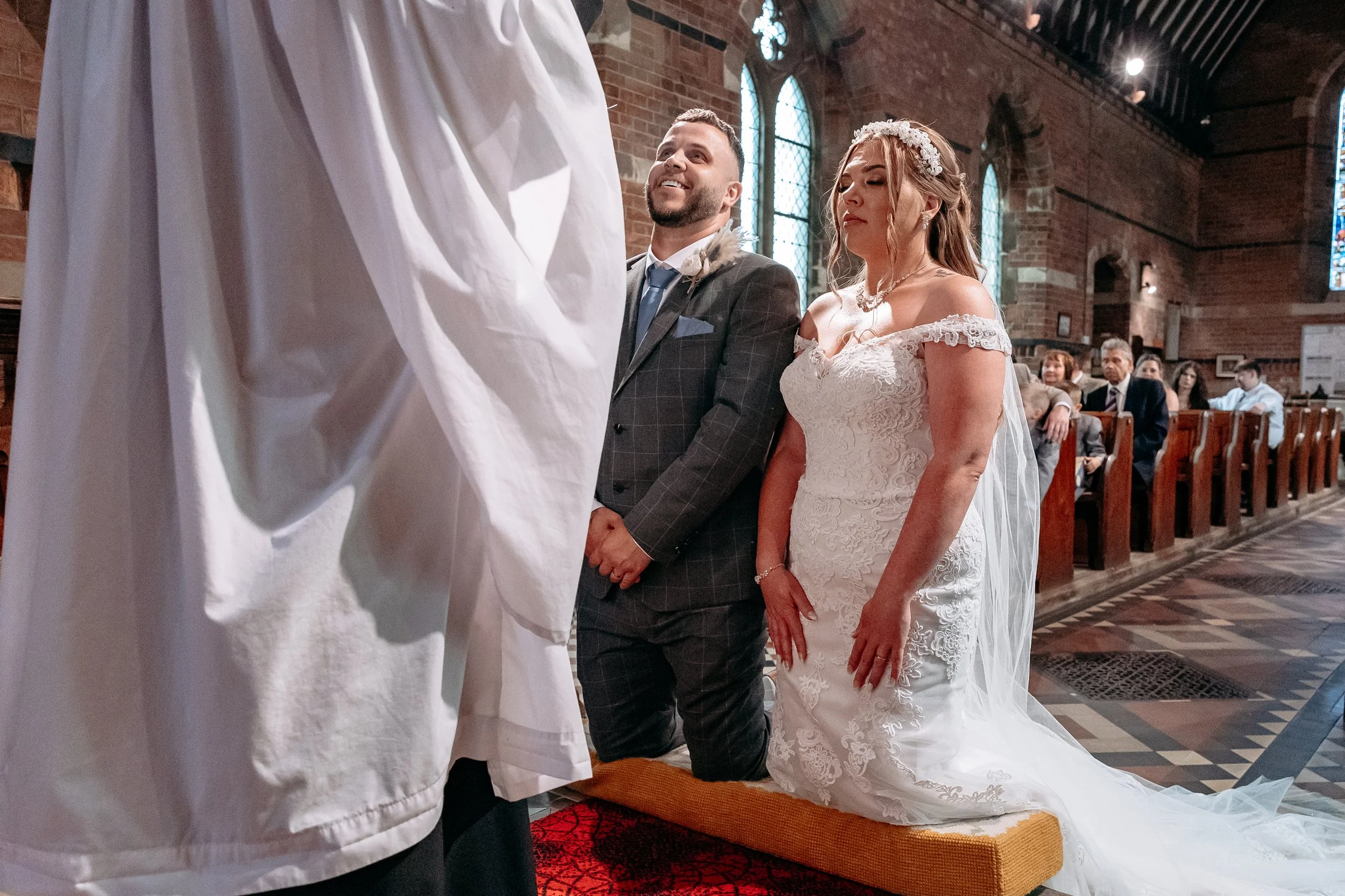 A bride and groom kneeling on a church pew during a wedding ceremony, facing the priest. The bride is in a white lace wedding gown with off-shoulder sleeves, and the groom is in a dark checkered suit. The church has brick walls, stained glass windows