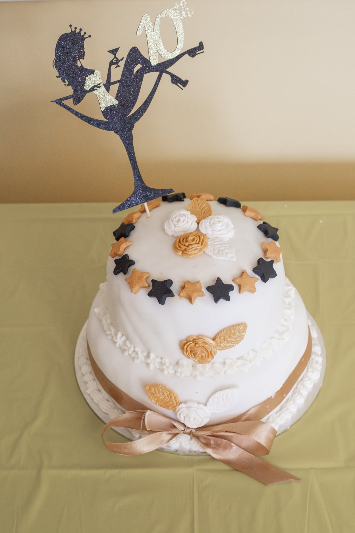 White two-tier birthday cake decorated with cream roses, gold and black star-shaped candies, and a beige satin ribbon around the base. A glittery cake topper features a woman balancing on a cocktail glass with the number "10" above her head.