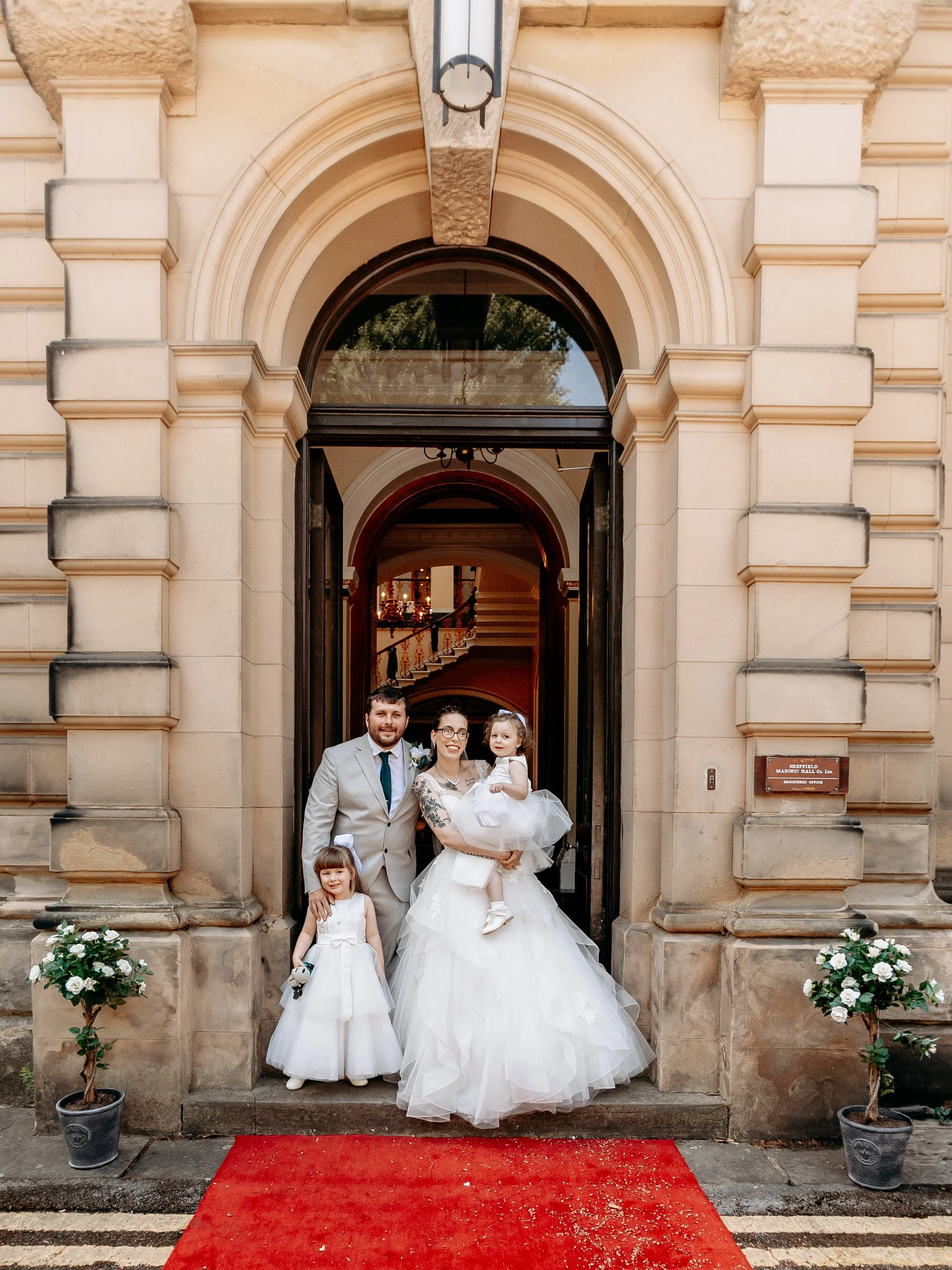 A wedding couple with two young girls standing in the doorway of a historic building. The bride is in a white wedding dress holding a young girl in a white dress. The groom is in a gray suit. Two potted white flower plants flank the doorway, and a re
