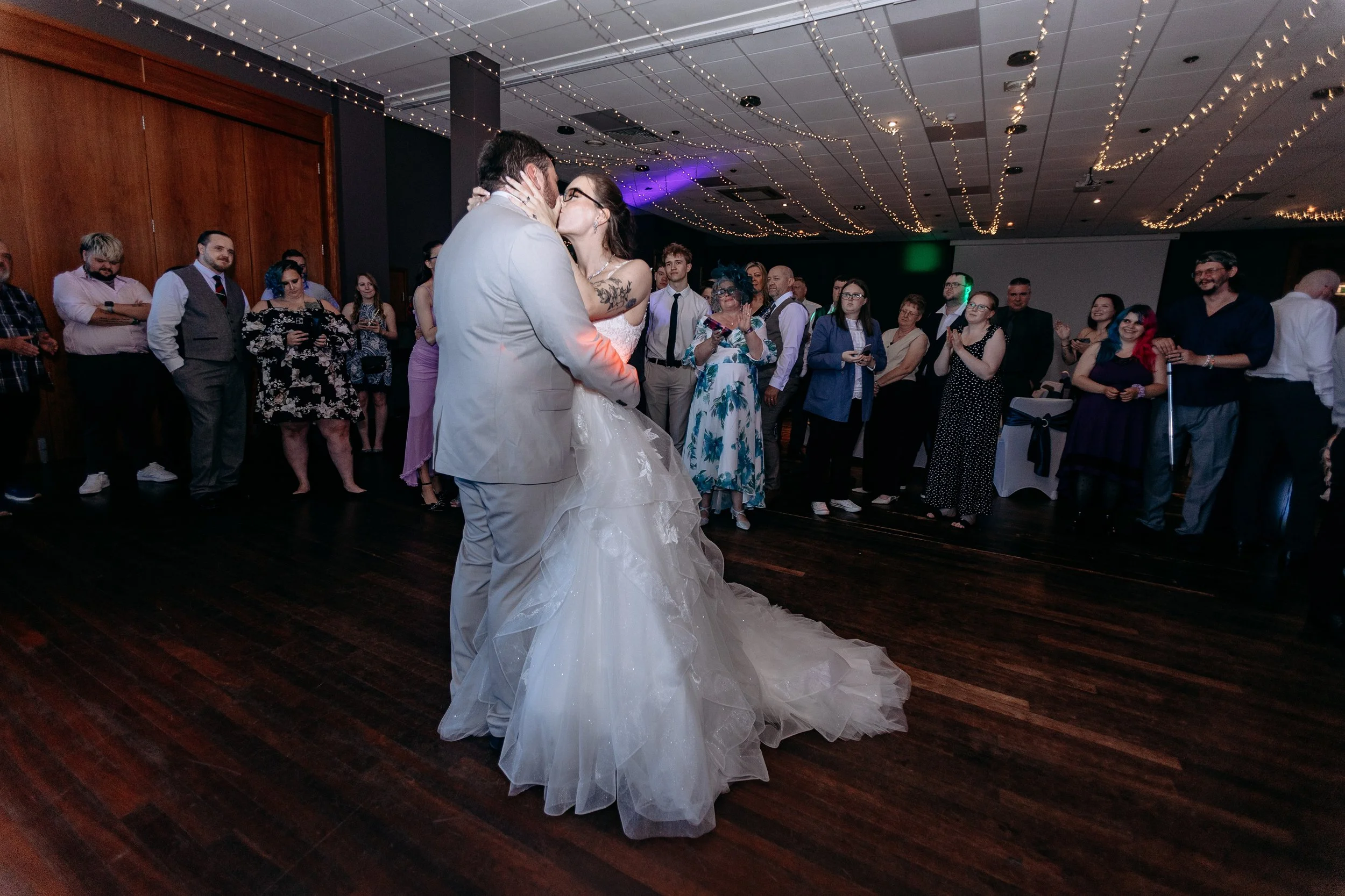 Couple in wedding attire sharing a kiss during their first dance at a wedding reception, with guests watching in the background under string lights.