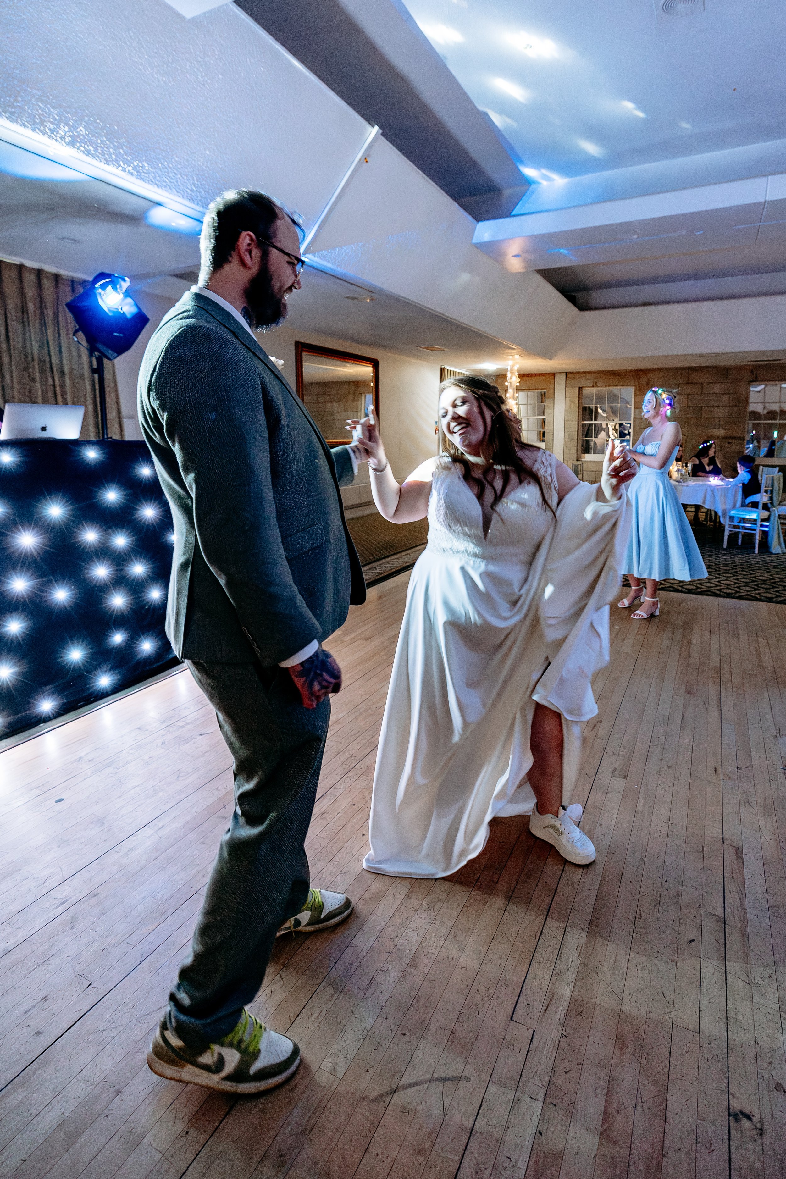 A man and woman dancing at a celebration, likely a wedding reception, on a wooden dance floor with other guests in the background.