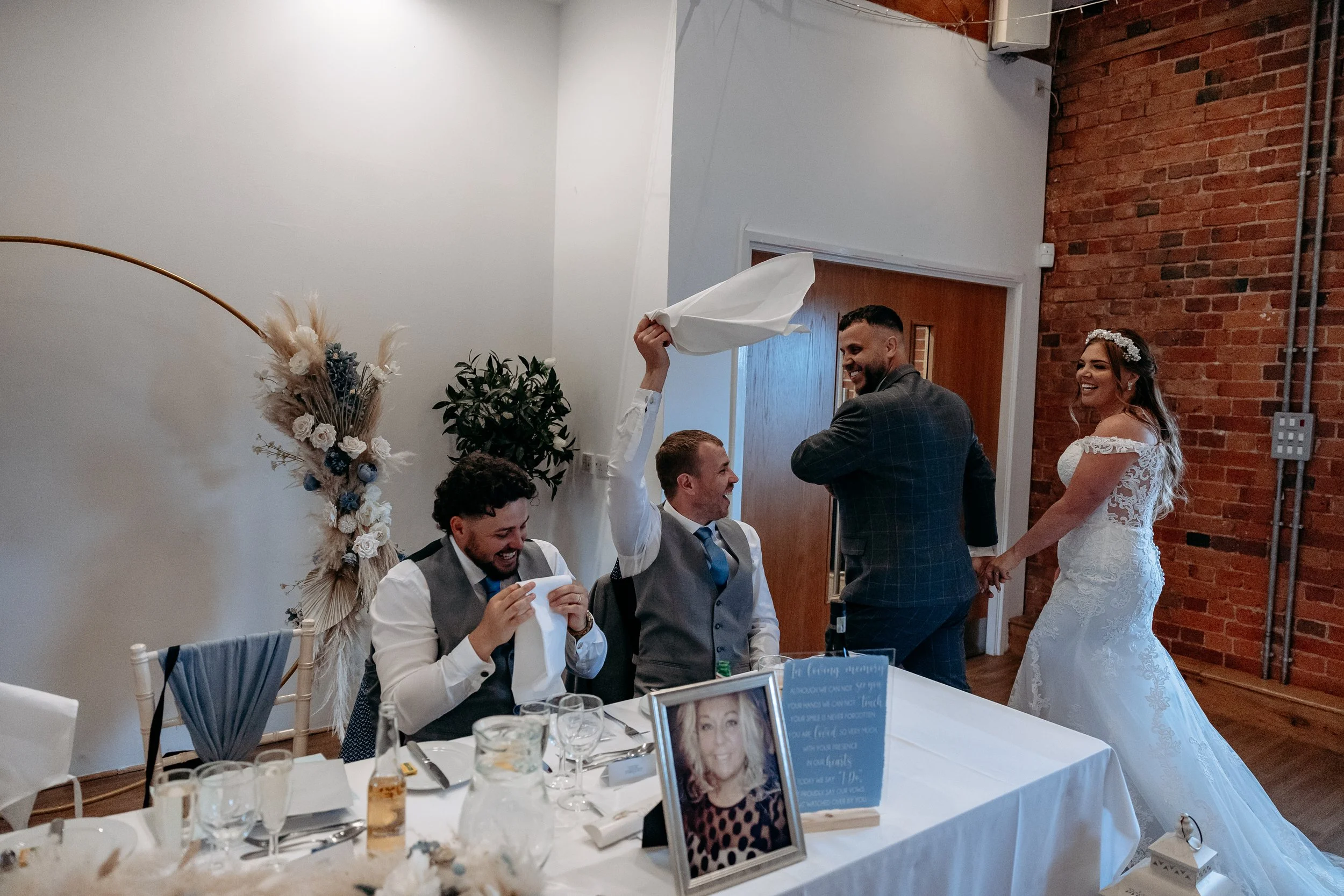 A bride and groom standing and holding hands at their wedding reception, with two groomsmen sitting at a table laughing, and a man standing off to the side, all inside a decorated room with a brick wall and floral decor.