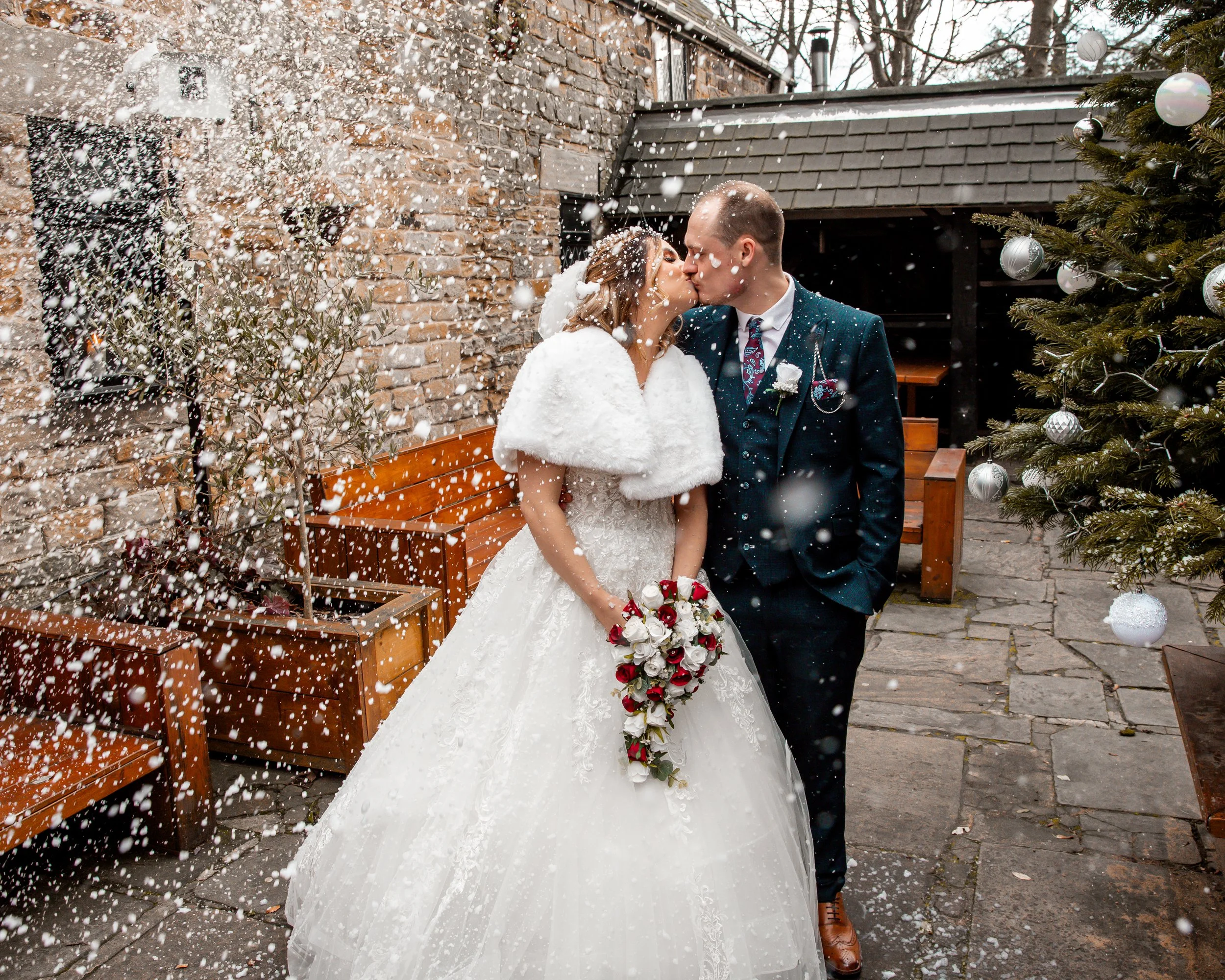 A bride and groom sharing a kiss outdoors in winter, surrounded by snow, a Christmas tree, and wooden benches, during their wedding celebration.